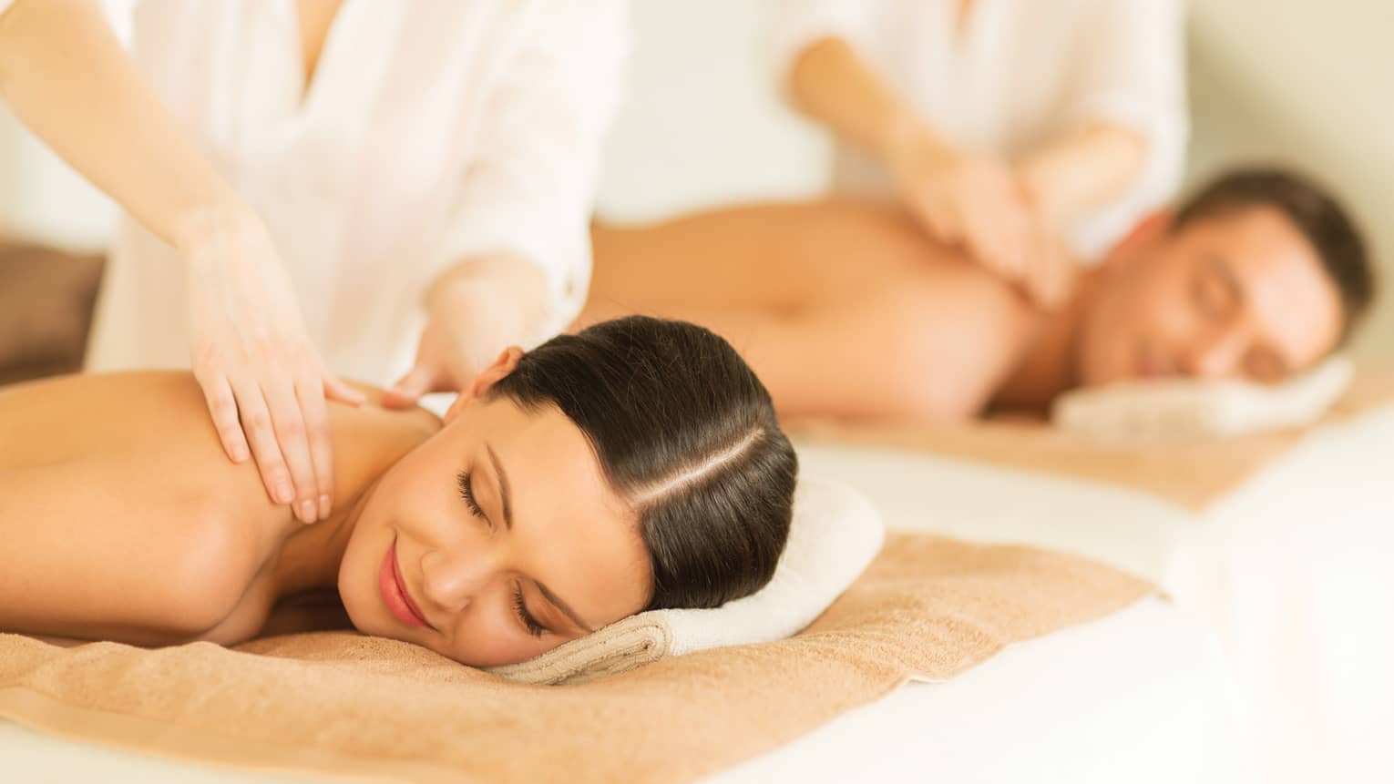 Woman and man lie side-by-side on massage tables as spa staff massage their bare shoulders