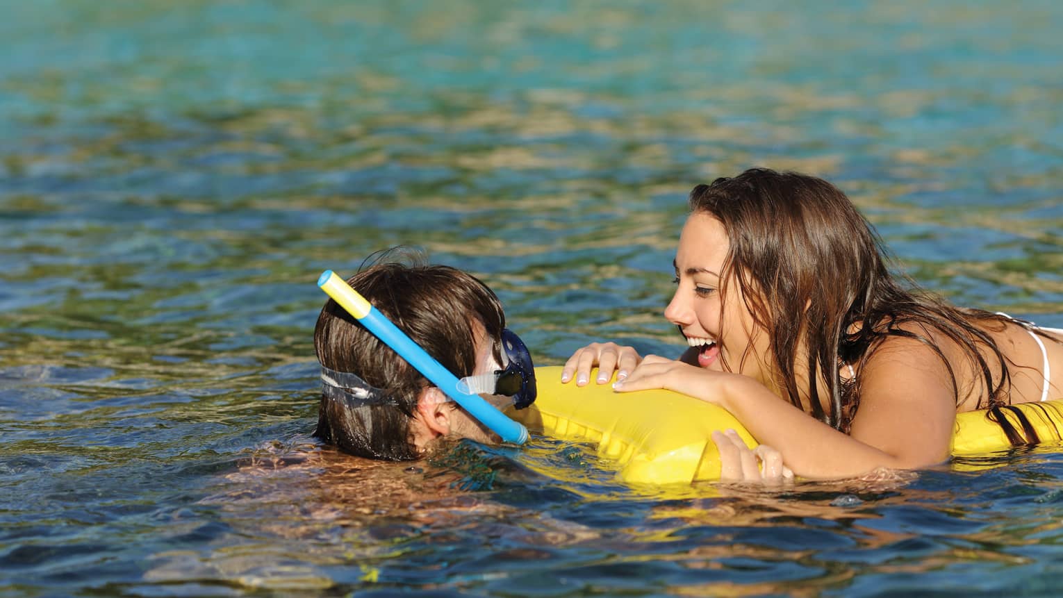 Child with snorkelling mask in water as woman floats on inflatable mat