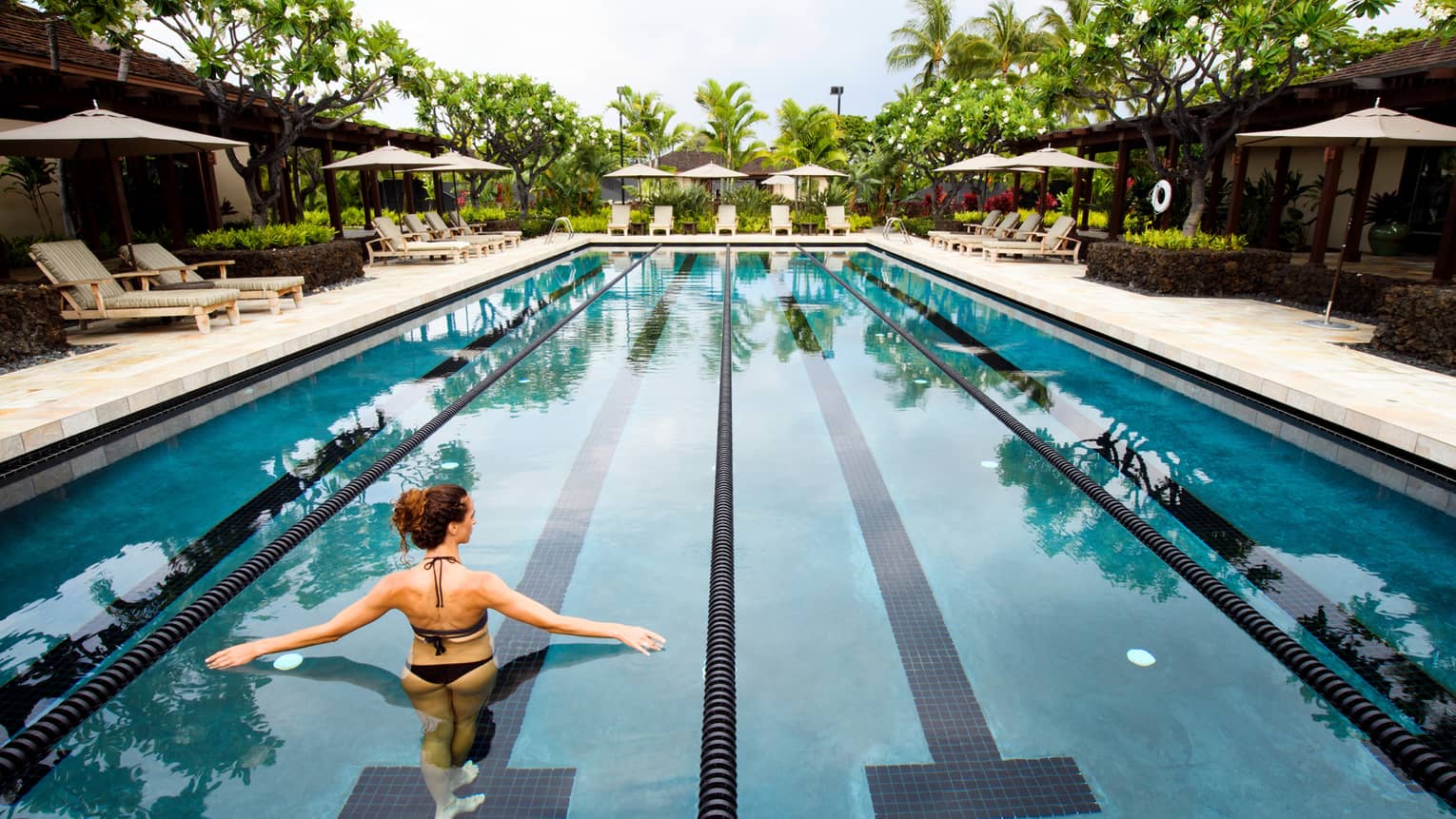 Back of woman in swimsuit wading in long outdoor swimming pool, arms outstretched