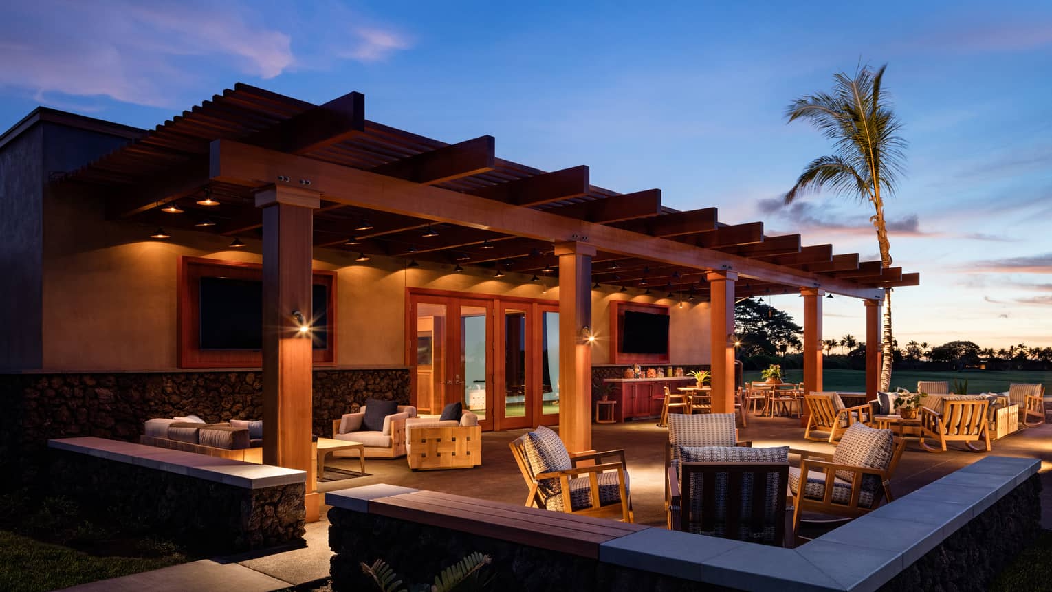 A warmly lit patio furnished with tables and cushioned armchairs under a clear, royal-blue night sky, sunset in the distance.