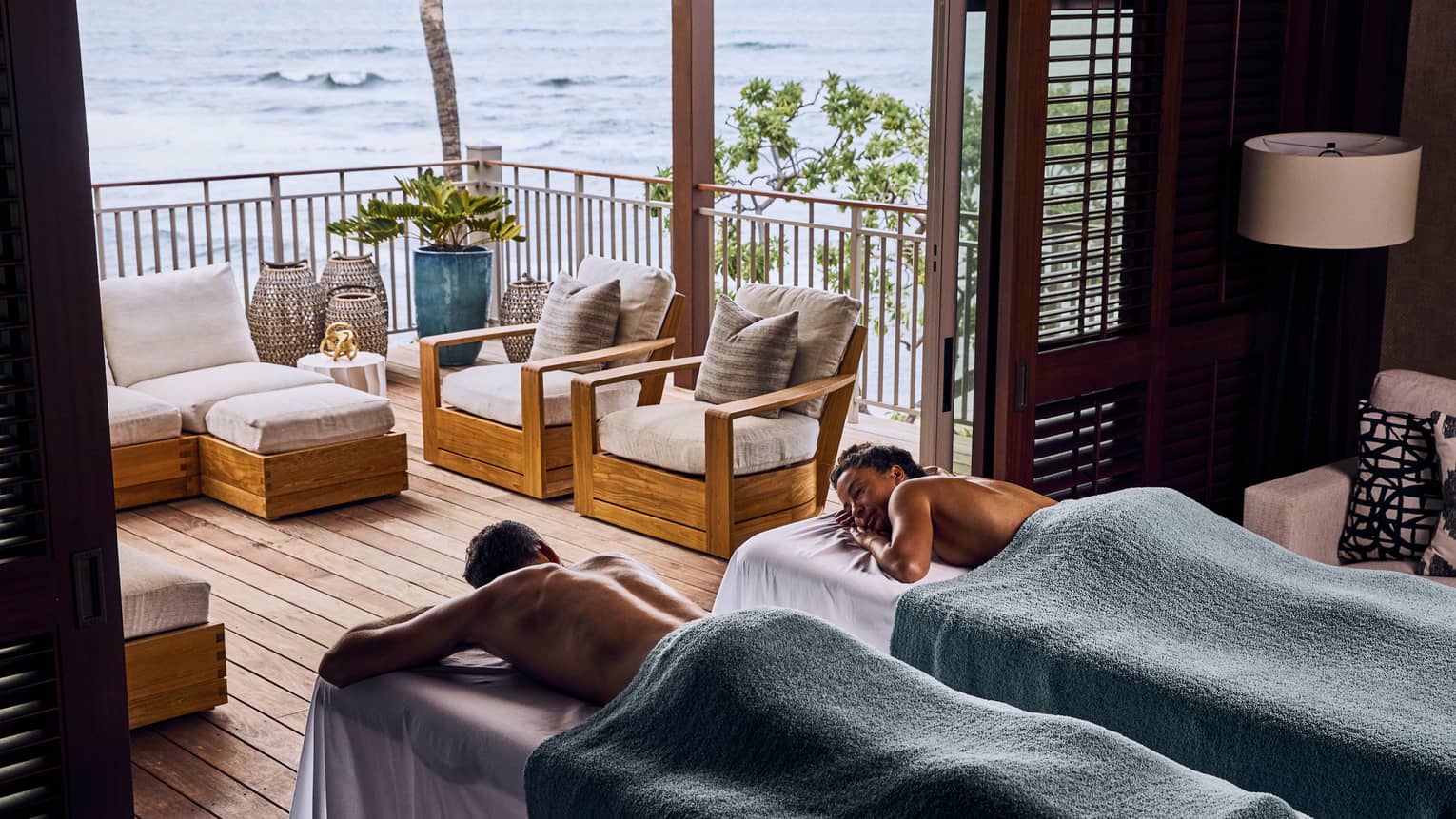 A couple covered in towels relax on massage tables in front of the open doorway of a balcony with an ocean view.