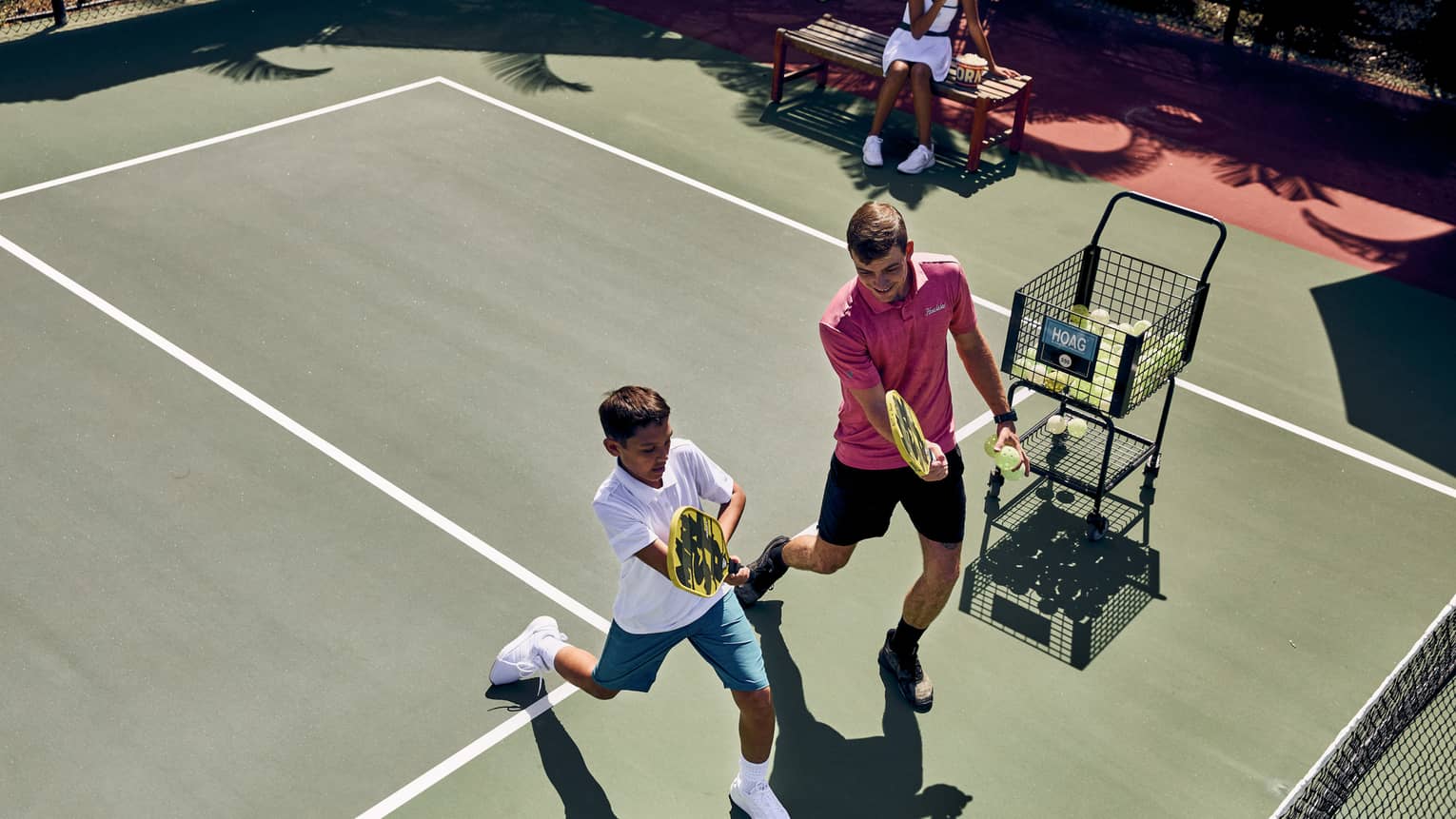 Aerial view of a pickleball pro instructing a player, both holding paddles while lunging forward, ball cart off to the side.