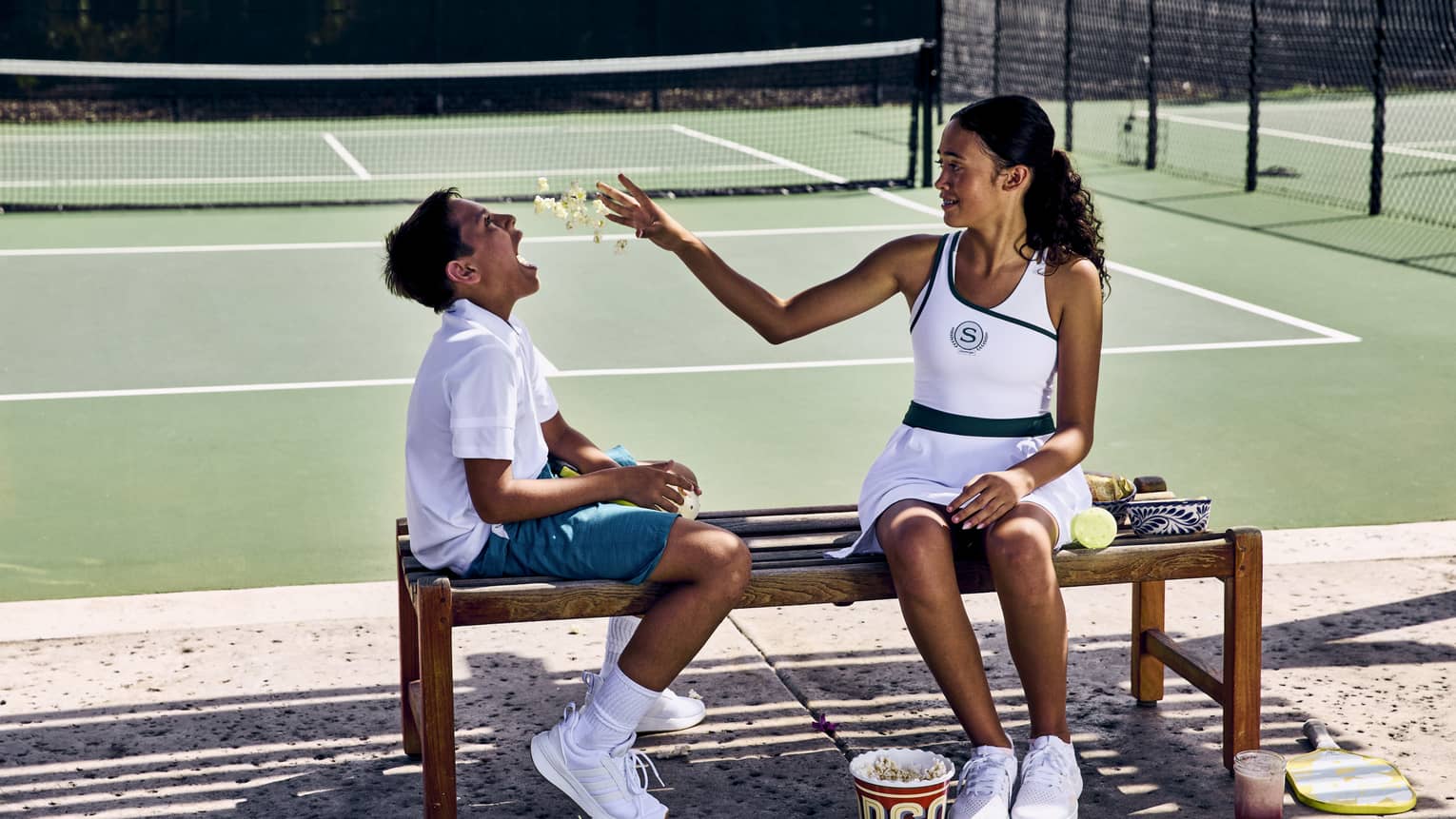 On a shaded wooden bench in front of a pickleball court, two children enjoy some popcorn, pickleballs strewn on the ground.