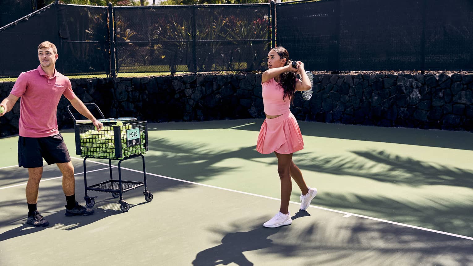 On a tennis court amid palm trees, a player finishes a swing over the shoulder; a pro stands by a ball cart, ball in hand.