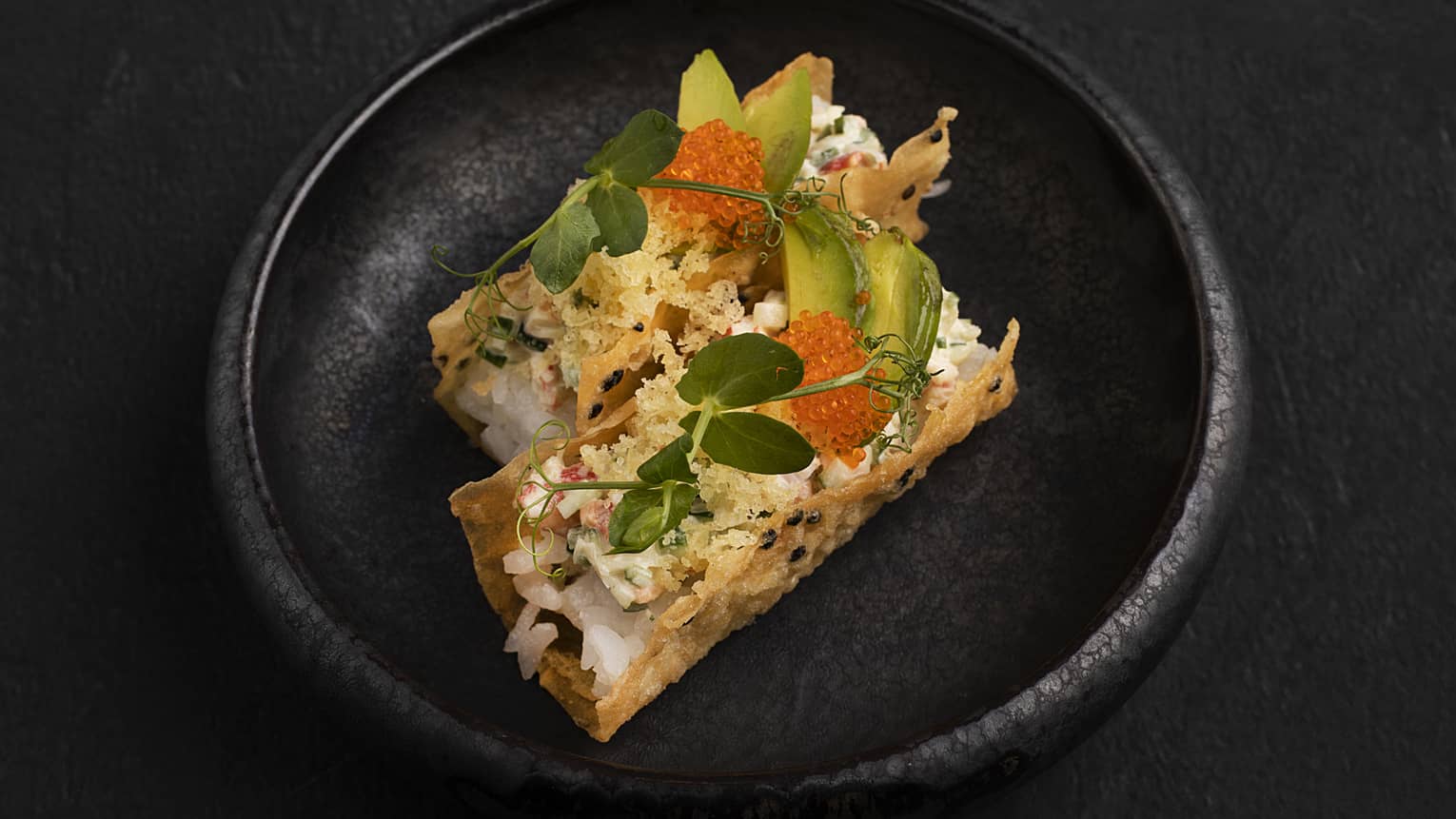 Two crispy shells filled with crab stick, cucumber, avocado, orange tobiko, tempura flakes and wasabi mayonnaise served on a black round plate