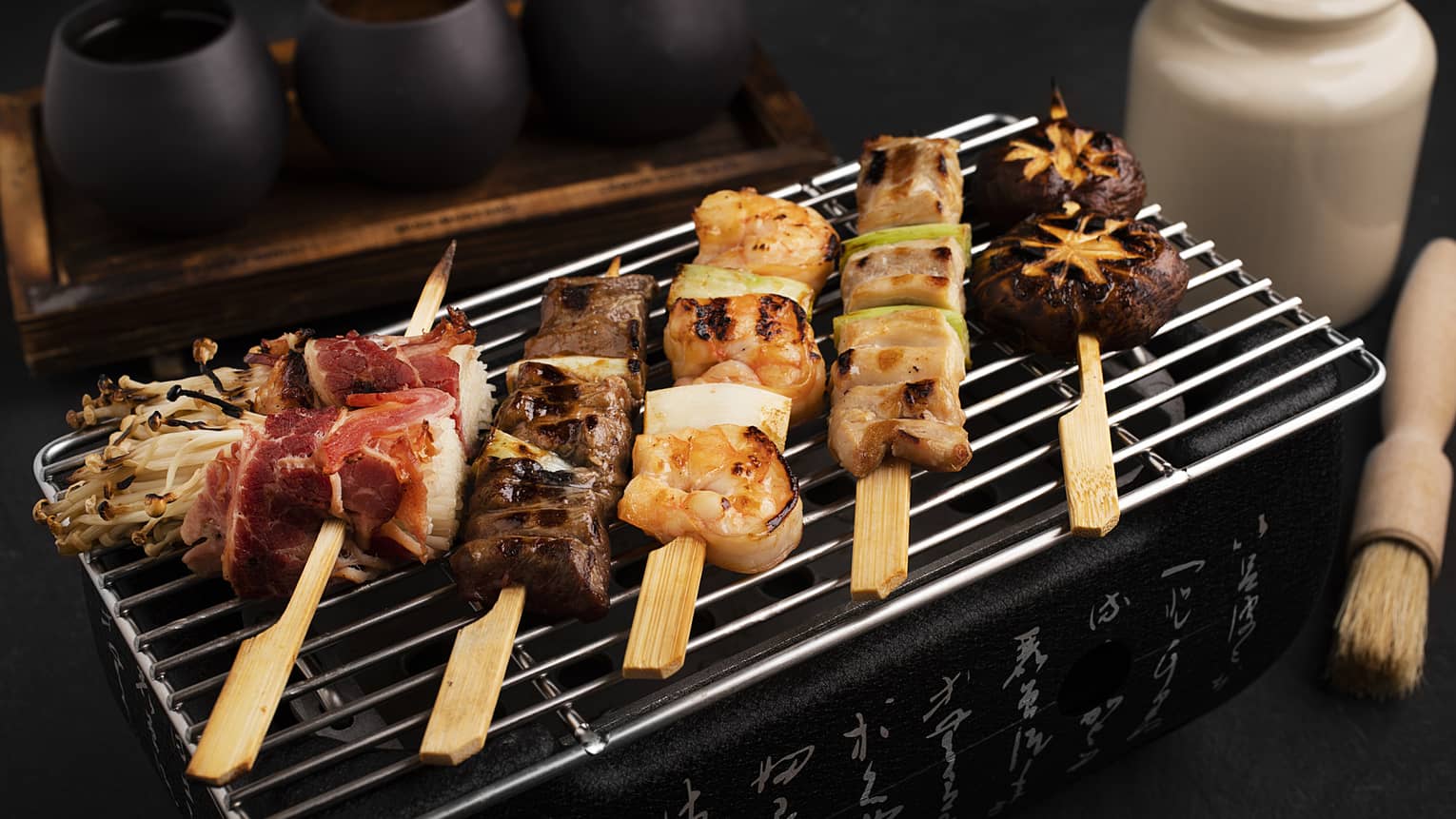 Chicken negima, beef kushiyaki, enoki bacon wraps, prawn skewers and shiitake mushrooms all on skewers laying on a grill