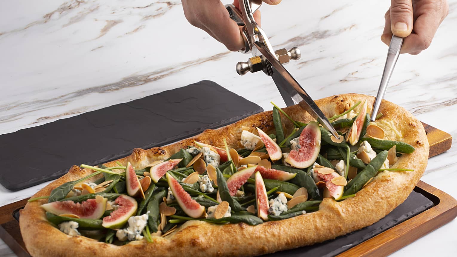 A large pizza topped with various fruits and vegetables, being cut into with a knife and pair of scissors.