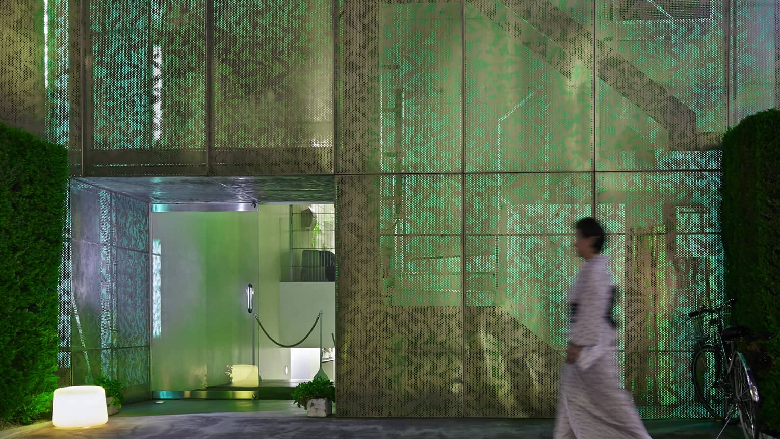 A person heads toward a door to a modern room, passing a multi-paned screened wall with a leaf pattern illuminated in green.
