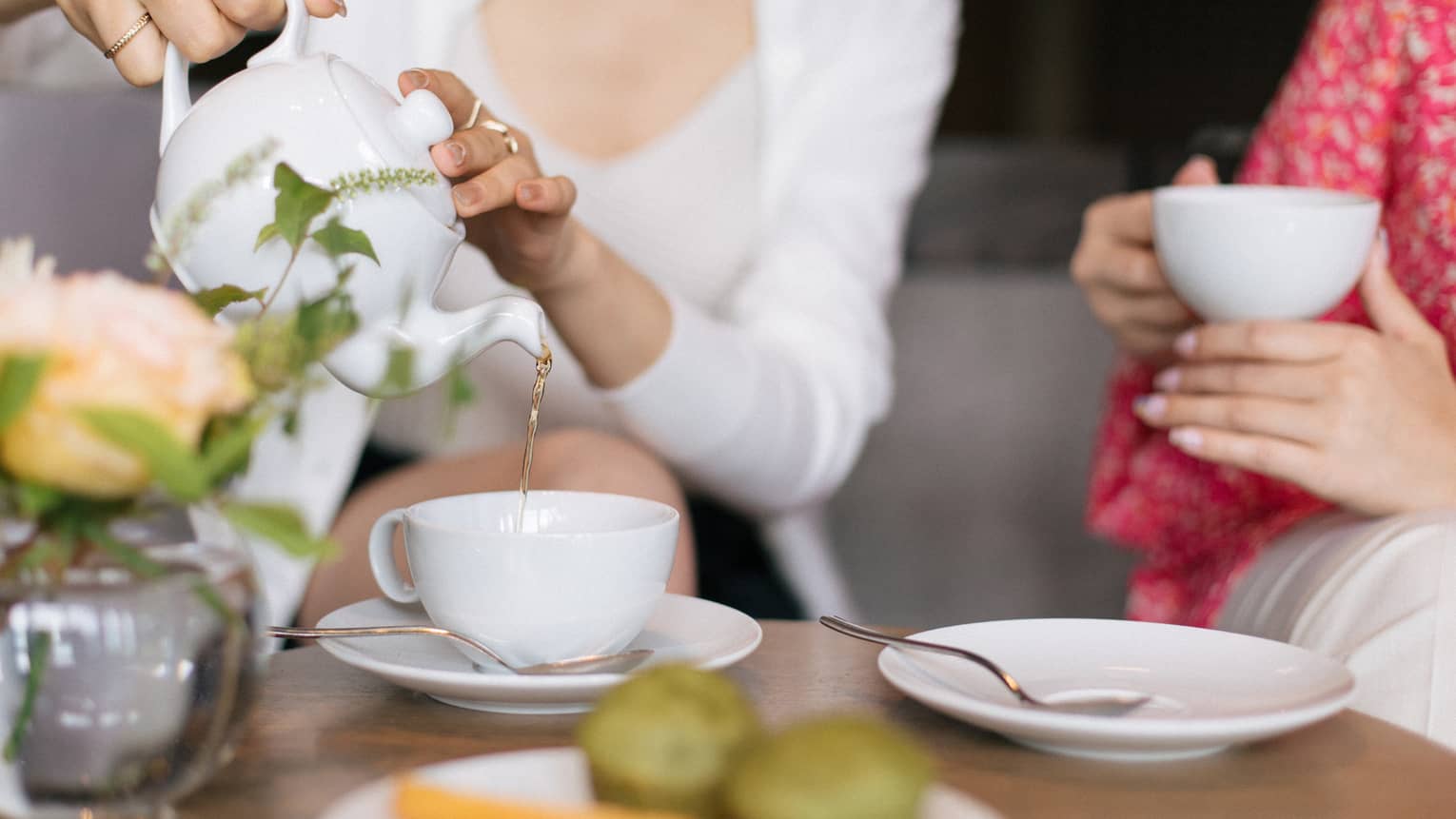 Two people sit at a table set for tea, with cups, saucers, a plate of small food items and a floral arrangement. One person is pouring tea from a teapot into one of the teacups.
