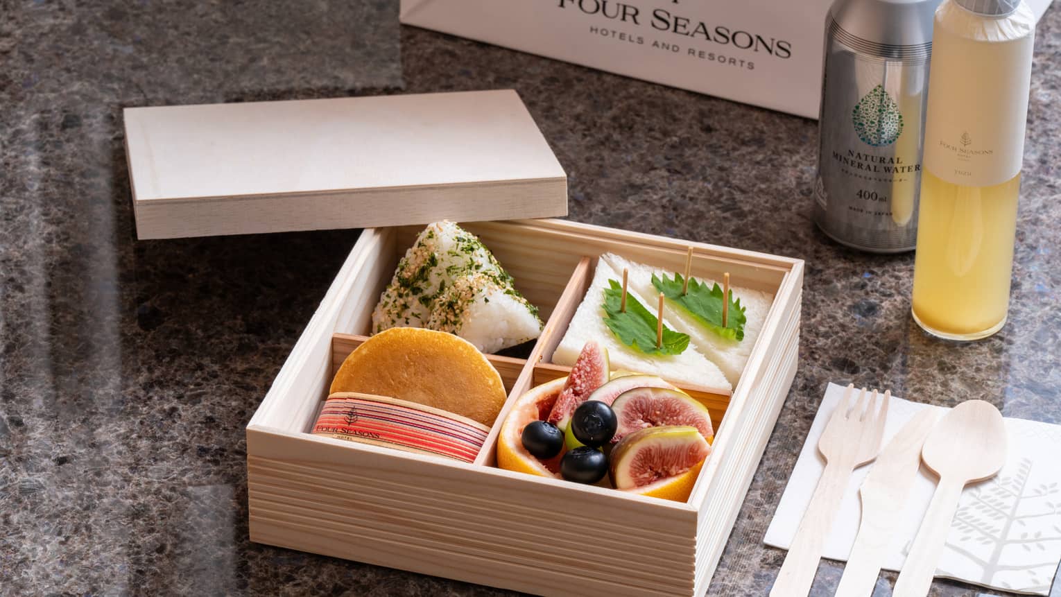 A wooden Four Seasons bento box with four compartments holding sandwiches, fruits and delicacies, beverage bottles nearby.