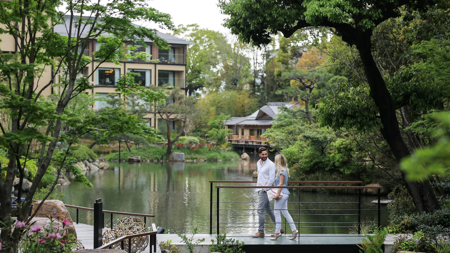A couple leisurely walks across a footbridge, engaged in a conversation, overlooking a tranquil pond surrounded by lush greenery in a park.