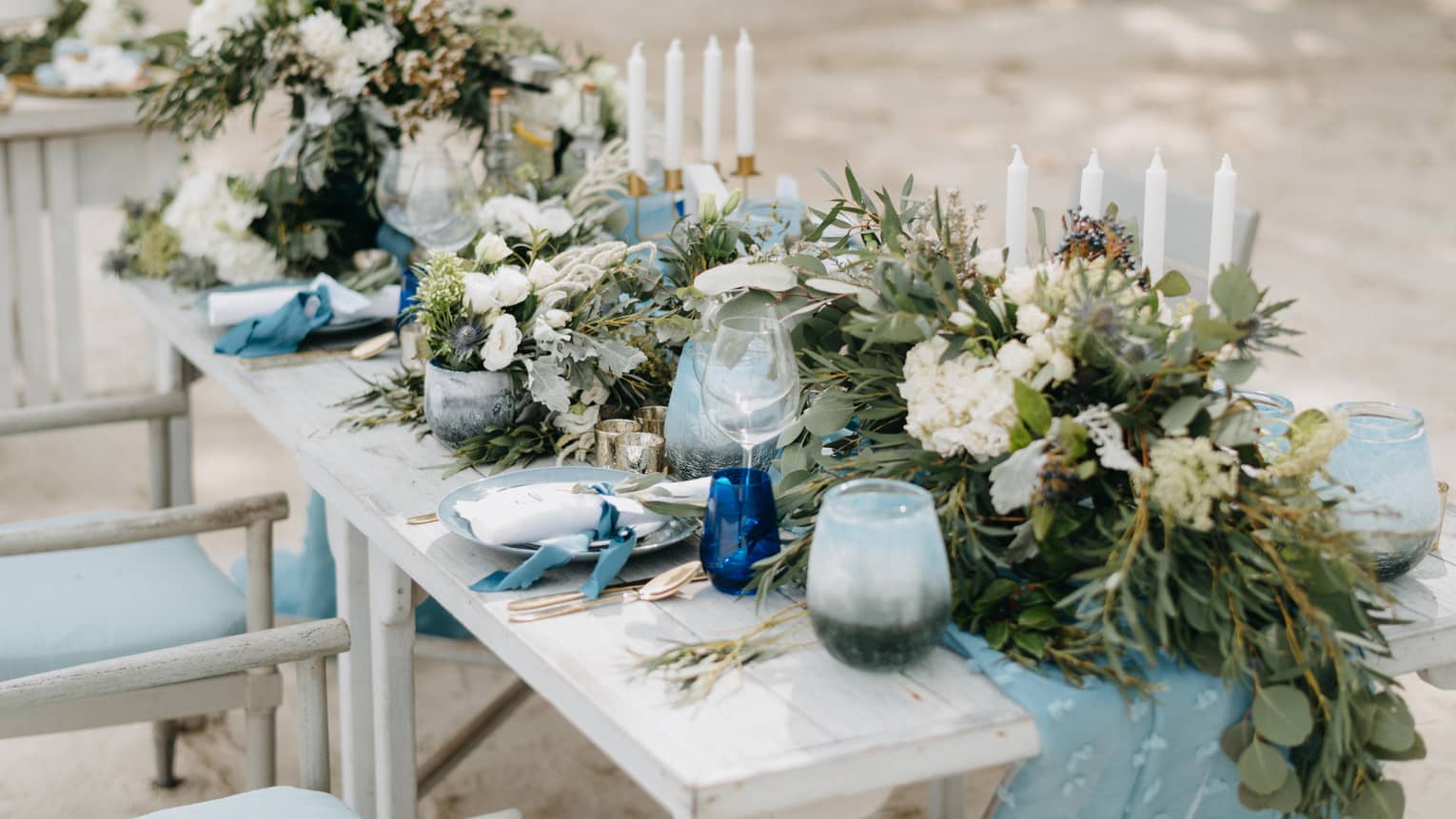 Outdoor rectangular table set for two and decorated with blue accents, green foliage and white tapered candles