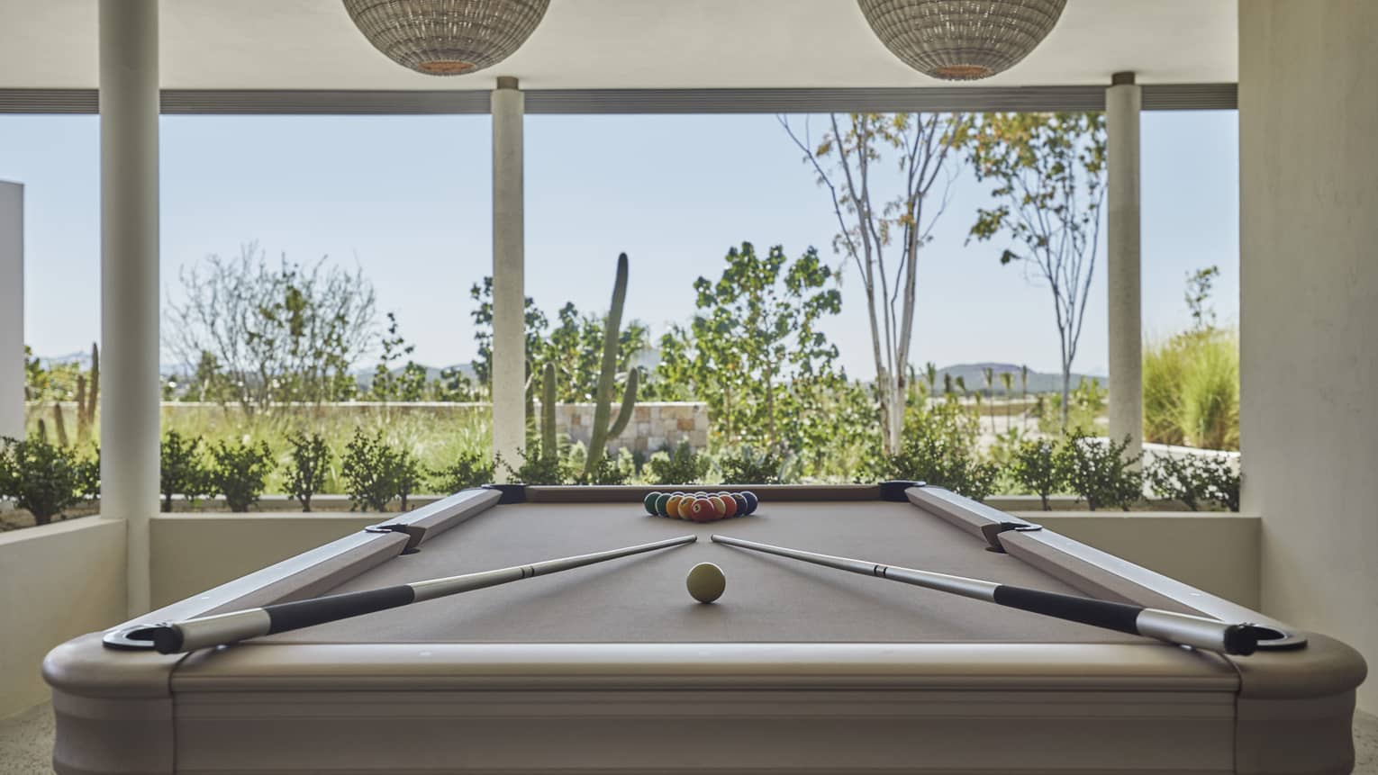 A billiards table next to large windows looking at grass and plants outside.