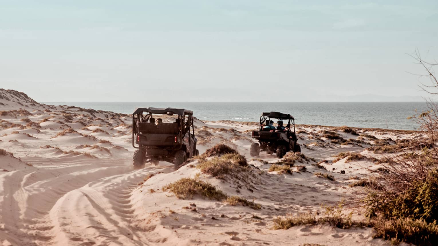 Two off road vehicles driving along dunes on a beach.