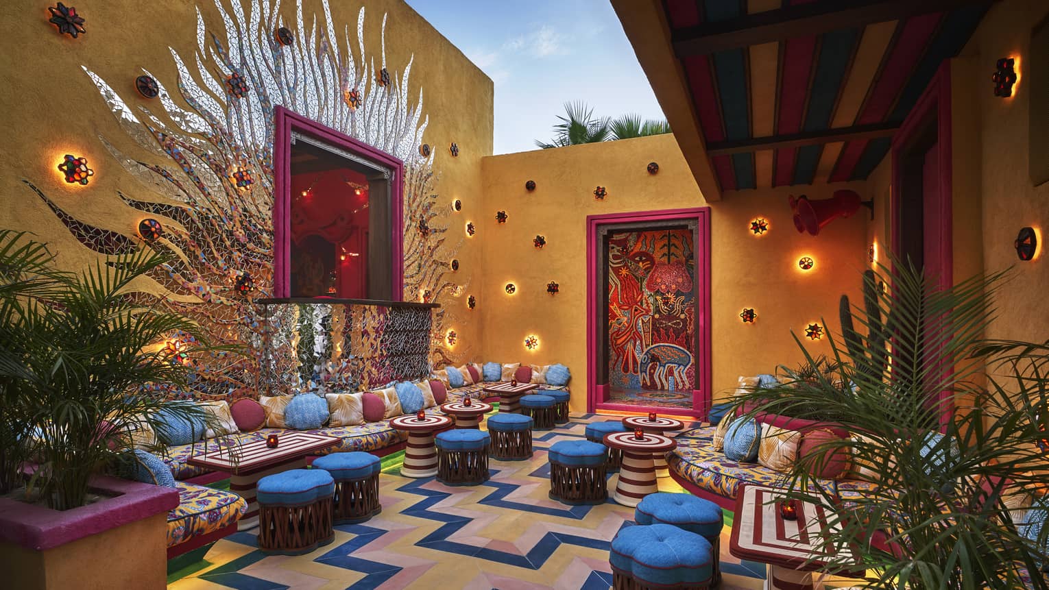 A colourful outdoor seating area with bright blue cushioned stools, colourful lights and plants.