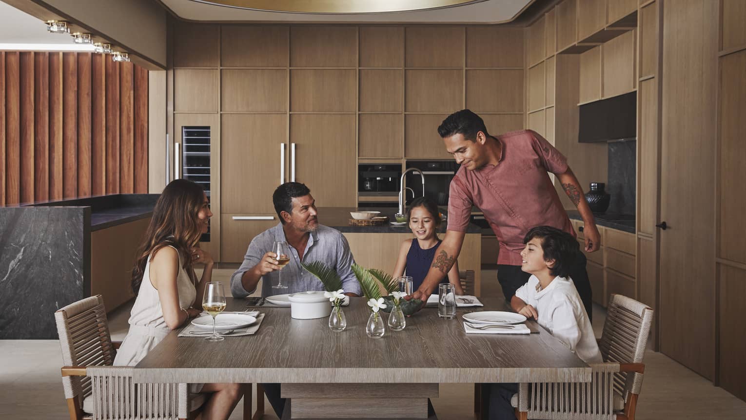 Four guests sitting at the table in a villa while a chef places a dish on the table.