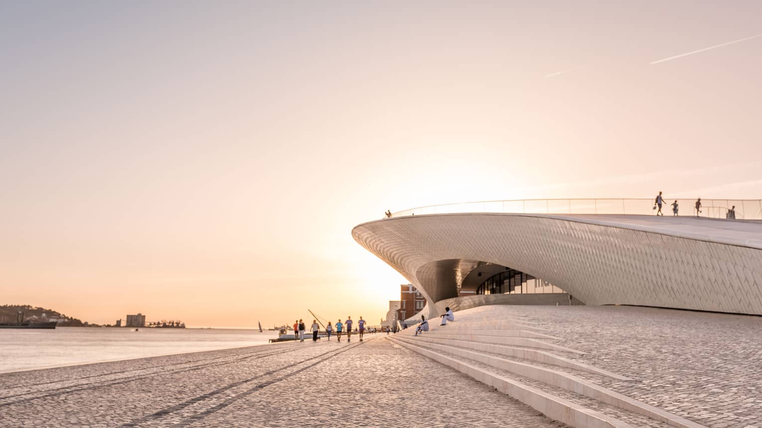 Tourists walk by a disc-shaped modern building next to a harbour. The setting sun reflects off the cobbled walkway. 
