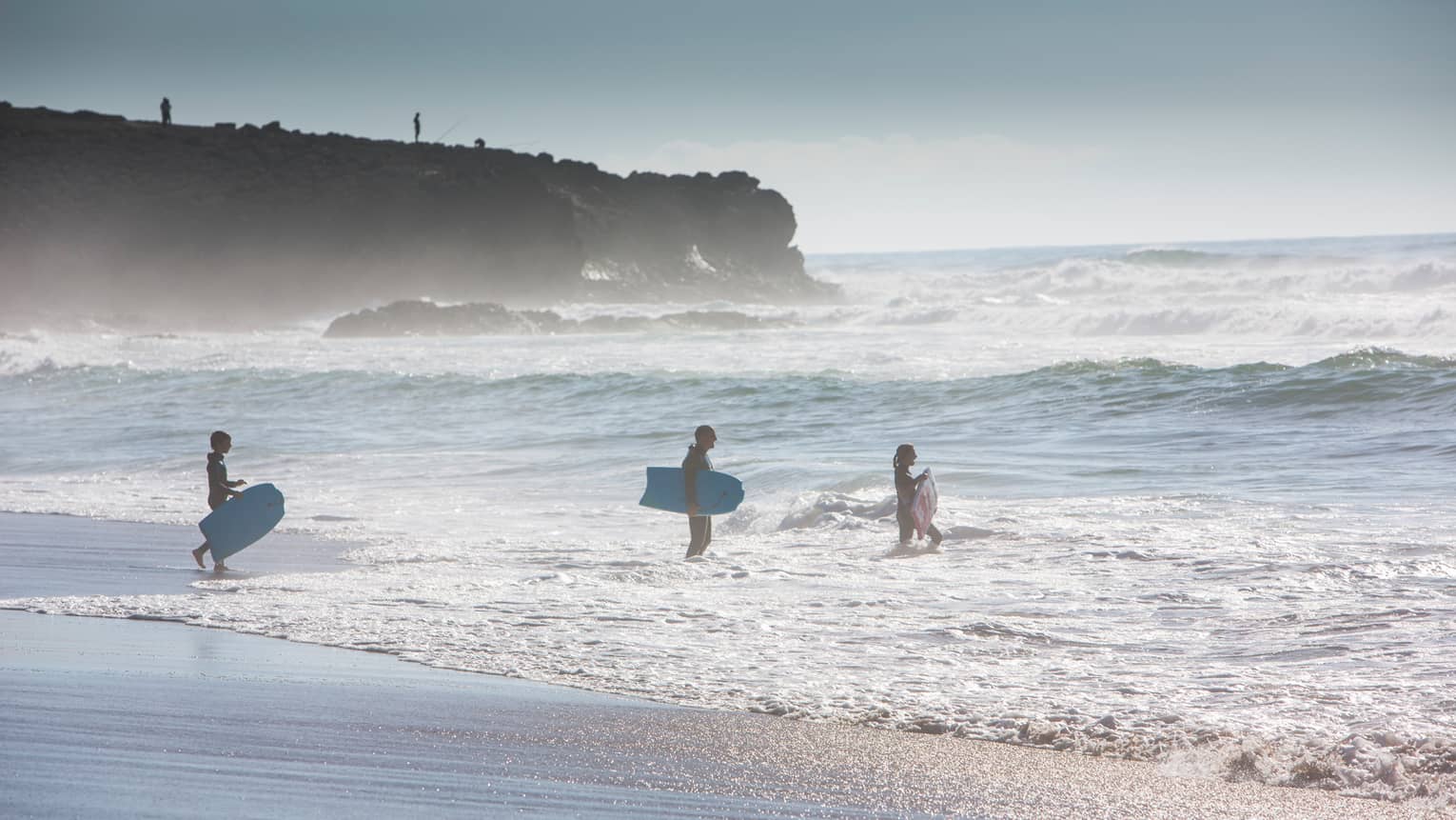 Silhouettes of man, woman and child all wearing wetsuits, holding blue surfboards, walking into ocean