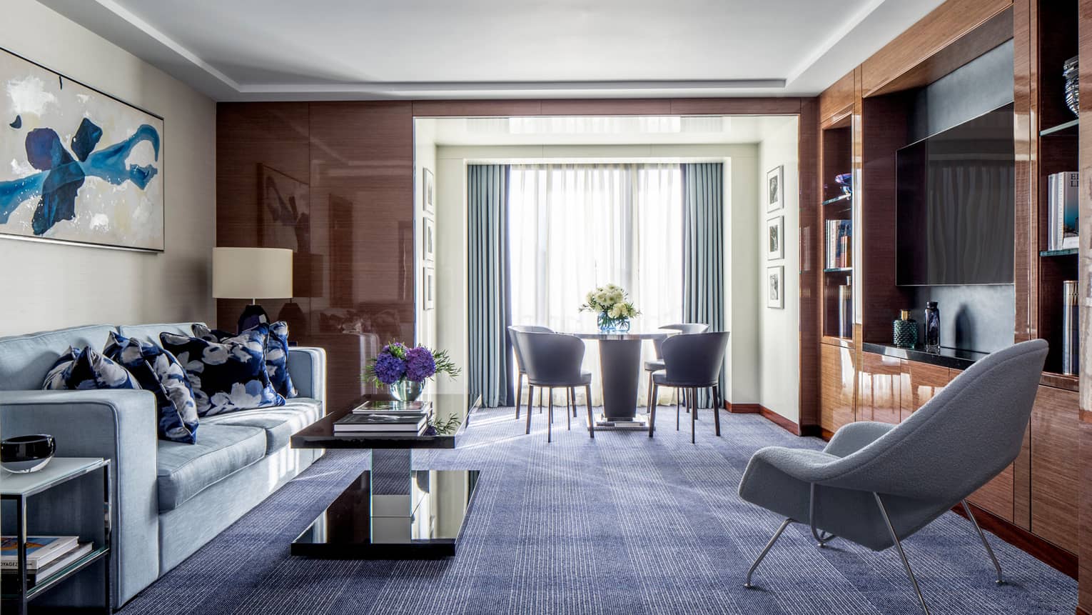 The Ambassador Suite at Four Seasons London Park Lane in Mayfair features a sophisticated and spacious living area. The room is elegantly appointed with rich wooden paneling and a deep blue carpet that contrasts beautifully with the light grey modern sofa and chairs. A large abstract blue butterfly painting dominates one wall, adding a vibrant splash of color. The space includes a sleek, dark coffee table and a dining area in the background with a round table and chairs, positioned near a bright window draped with sheer curtains.