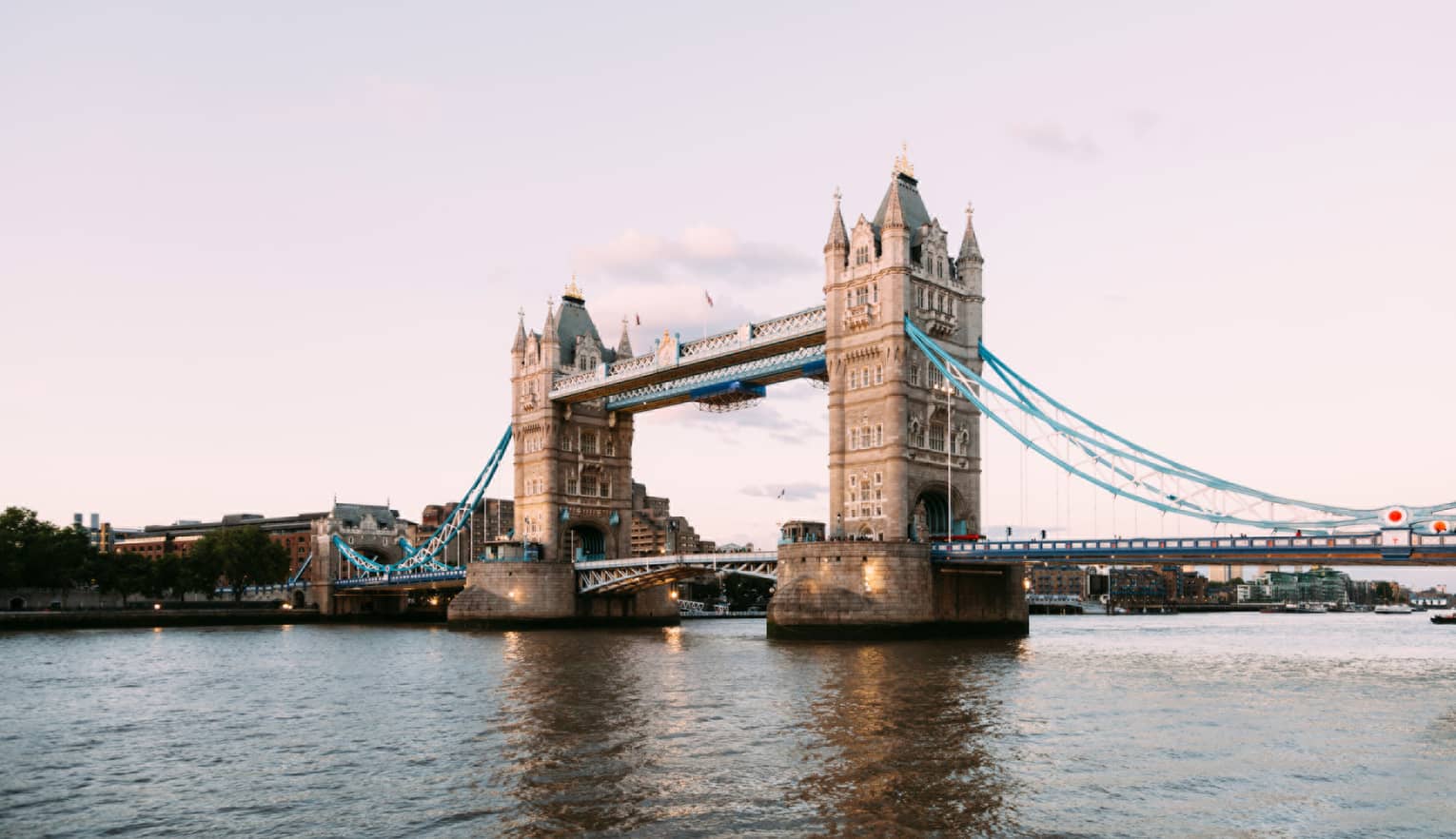 The iconic Tower Bridge of London spanning the dark Thames River in the foreground against a pale, dusky sky.