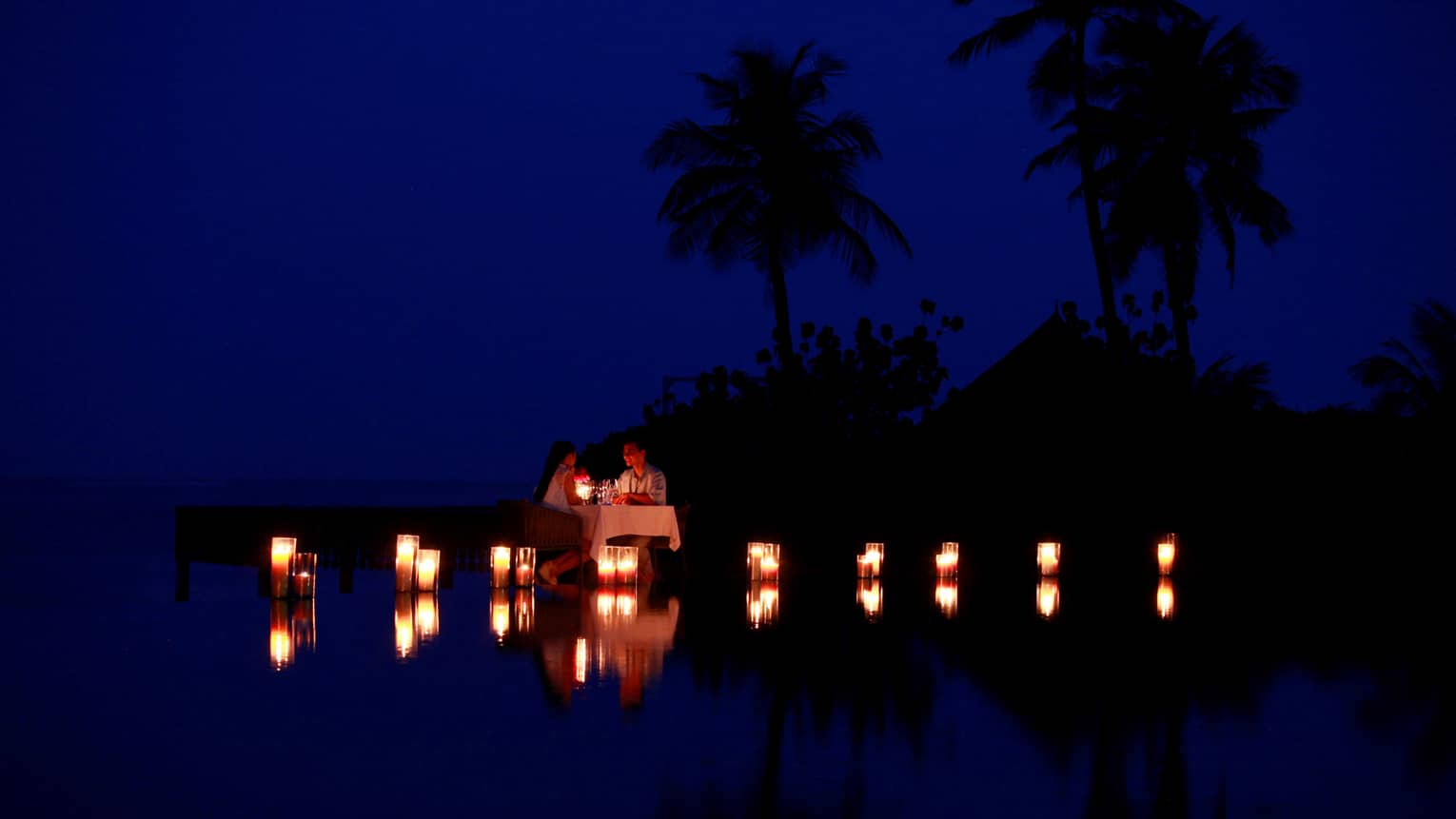 Couple dine by lagoon at small table surrounded by glowing candles at night