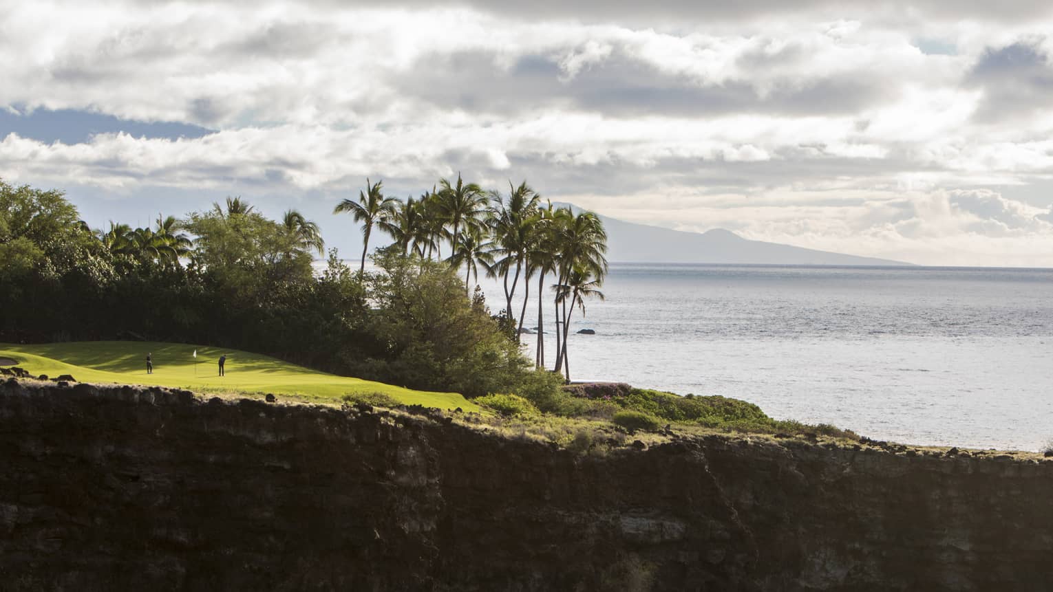 Long view of two people on clifftop golf green aside lush trees and towering palms; fluffy clouds, ocean and mountain beyond.