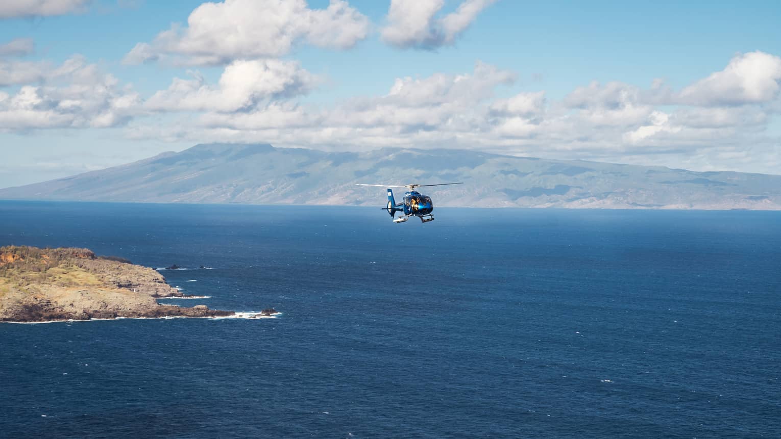 A helicopter flies over open ocean with a mountainous island and blue sky dotted with clouds in the background