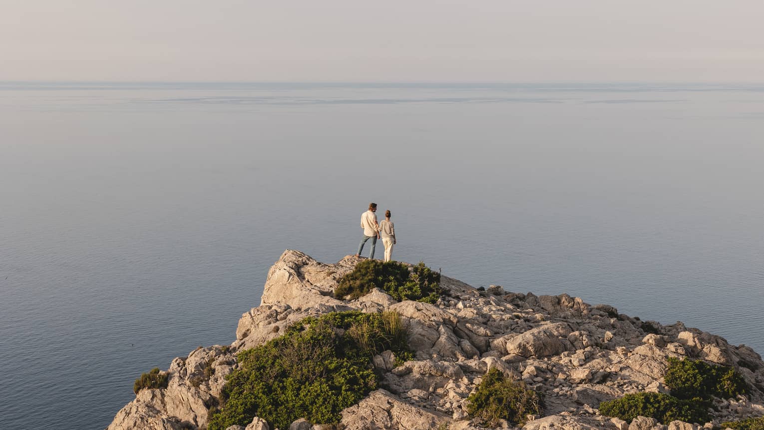 Distant view of a couple in hazy sunlight gazing at an expanse of tranquil water from atop a cliff dotted with shrubs.