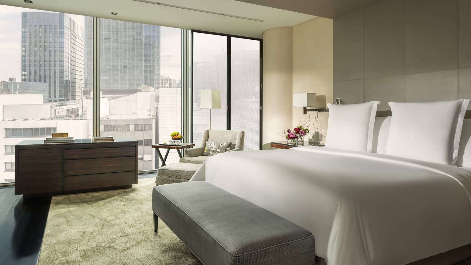 Modern hotel room with a king bed, grey tufted bench, cozy seating area, dresser and large windows offering city views