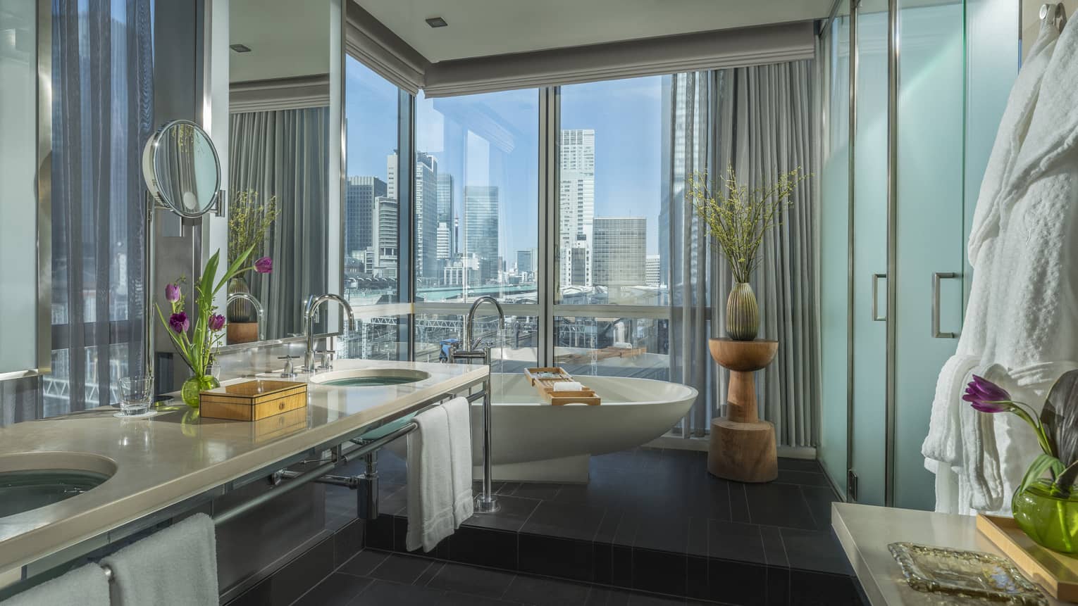 Spacious bathroom with a freestanding bathtub, double-sink vanity and floor-to-ceiling windows offering city views, decorated with flowers