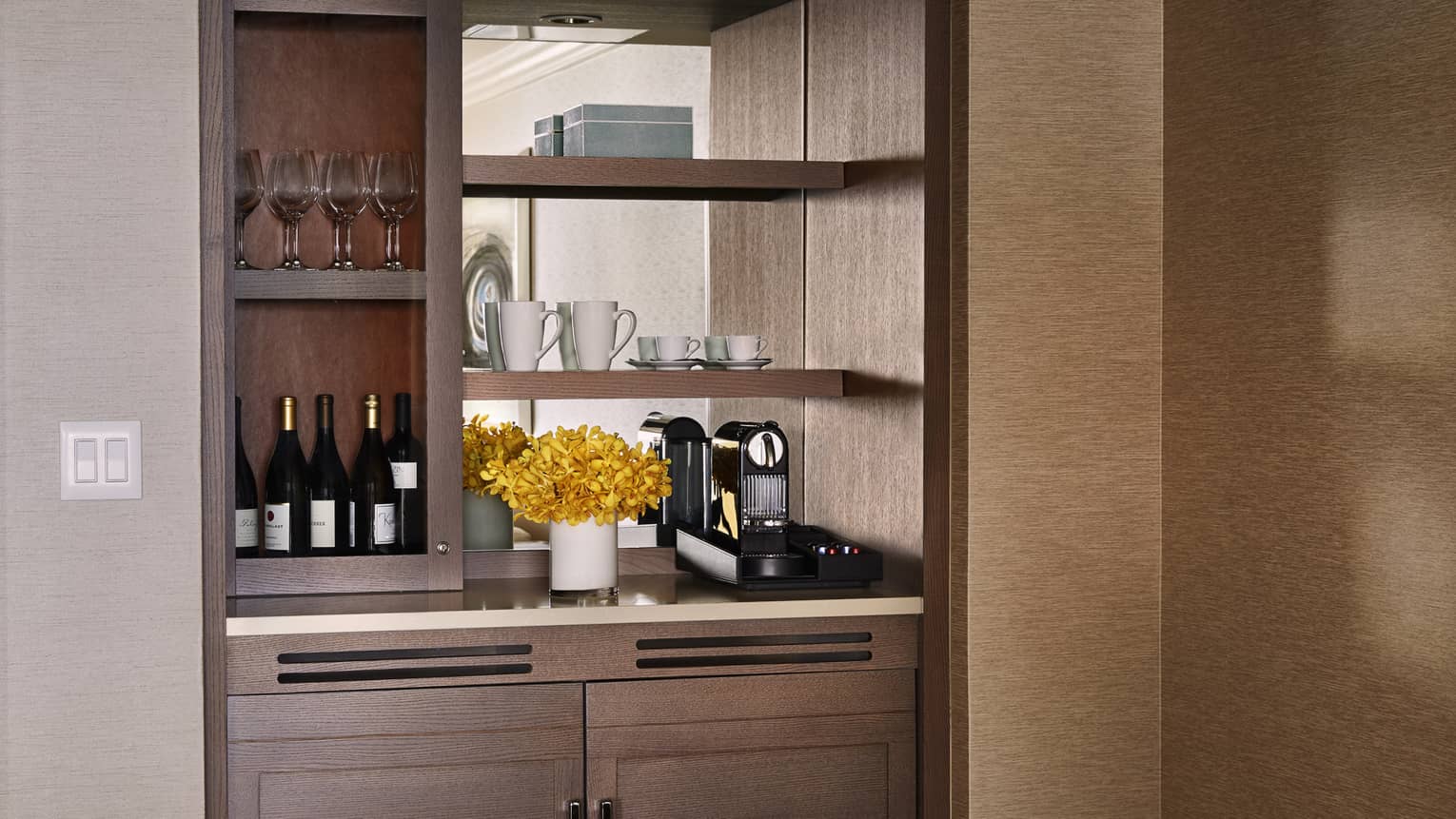 Resort suite mini bar with wine bottles, glassware, coffee maker and yellow flower arrangement on wooden shelves