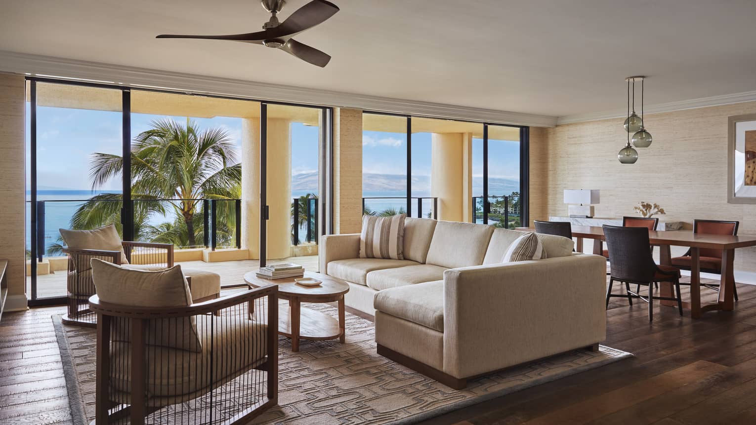 Spacious living and dining area with a beige sofa, armchairs and dining table, opening to a balcony with ocean and palm trees view at a luxury resort suite