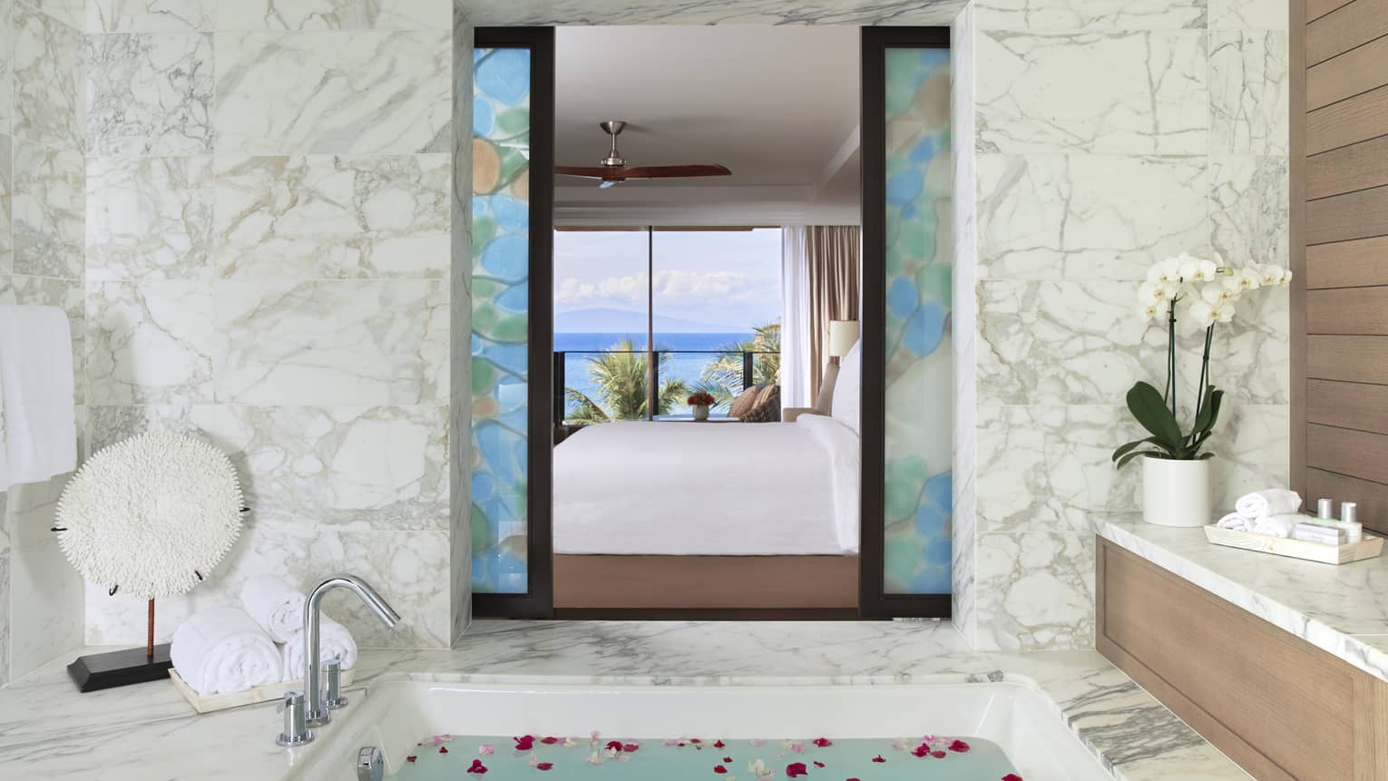 Luxurious bathroom with marble accents, a soaking tub with rose petals and a view of the bedroom and ocean through decorative glass panels