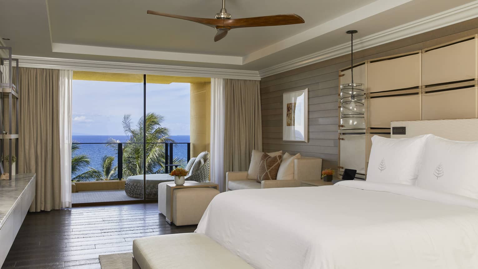 Modern bedroom with king bed, neutral décor, seating area and balcony overlooking the ocean and palm trees