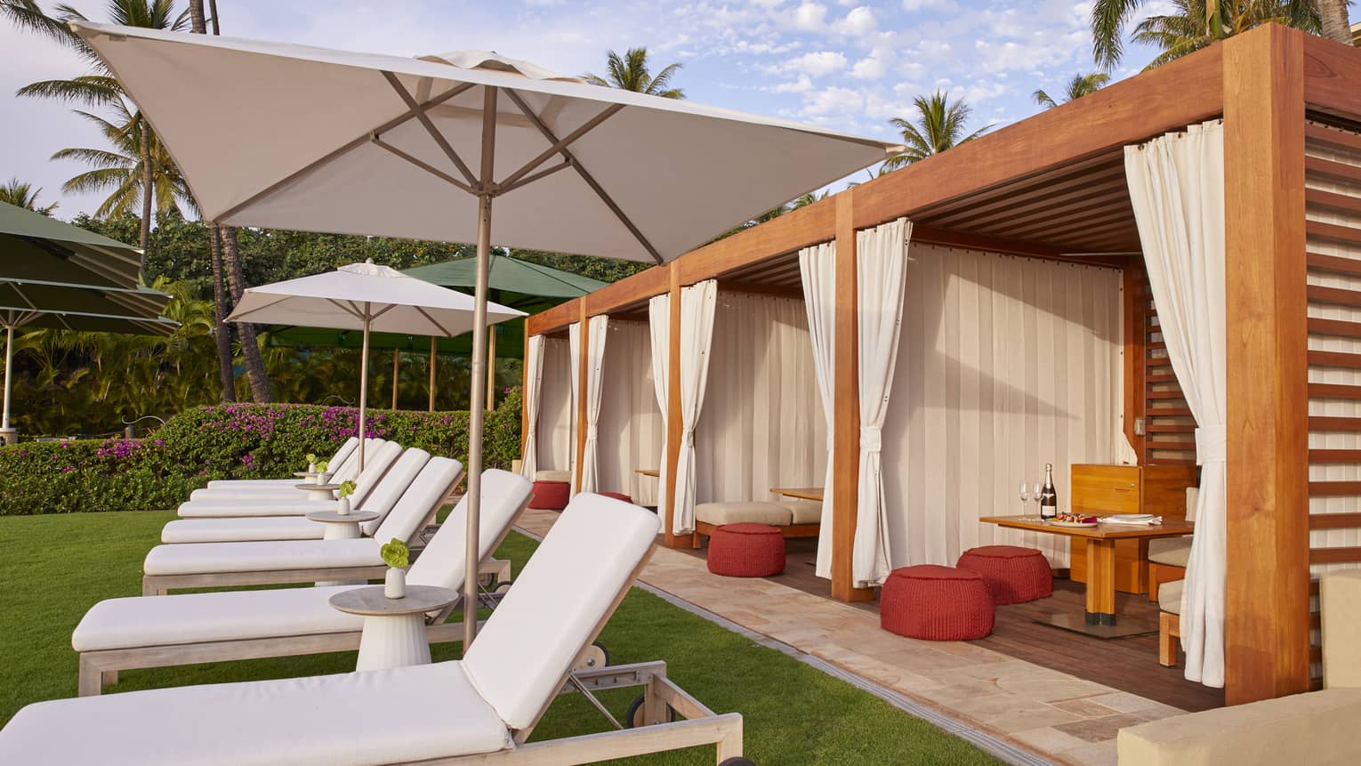 Patio lounge chairs with white cushions in front of three cabanas with curtains