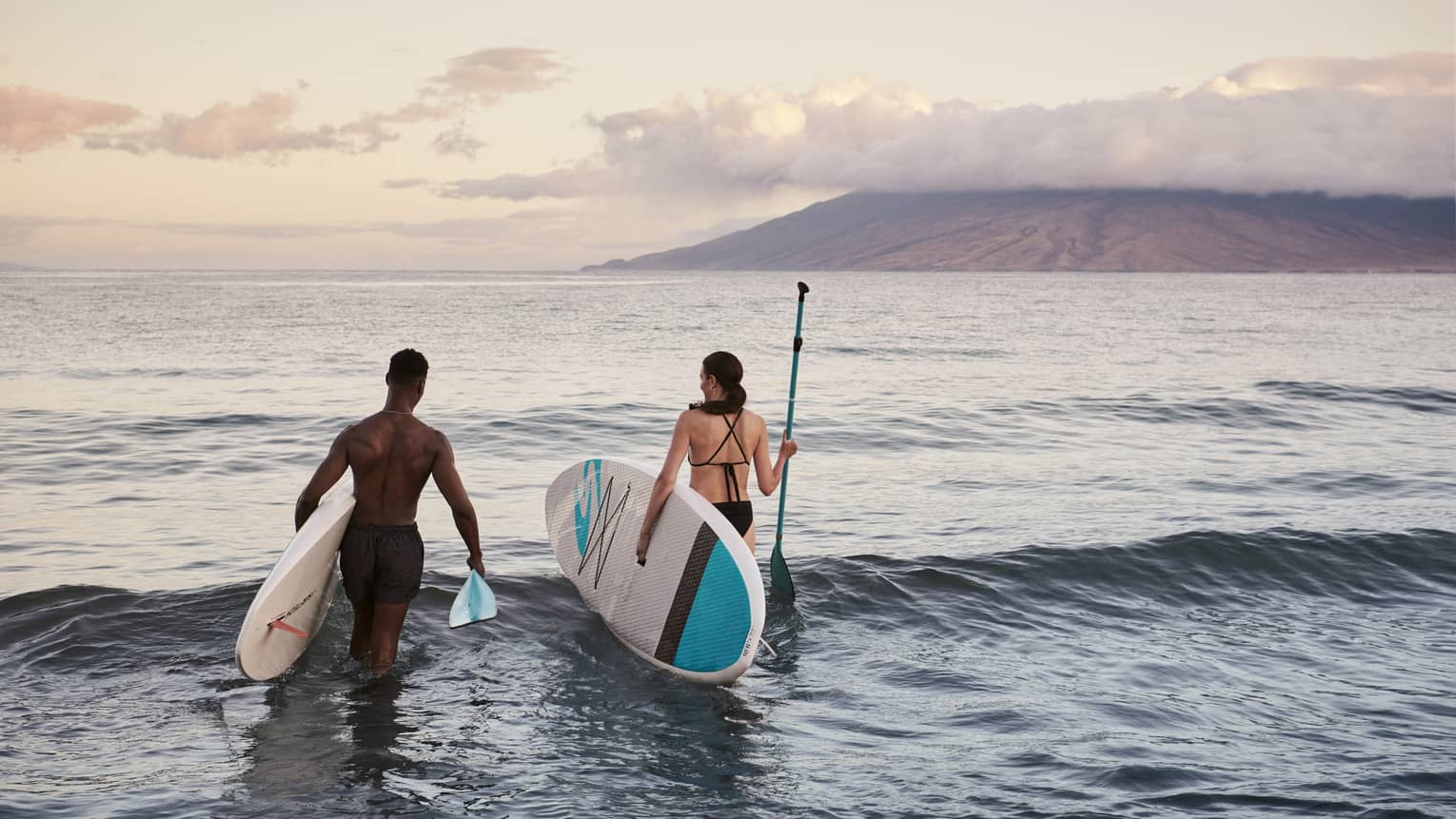 Two people standing in the ocean holding paddleboards with mountains in the background