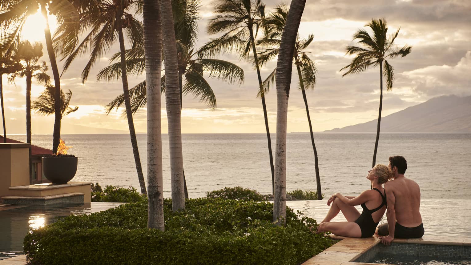 Couple relaxing by an infinity pool with palm trees, overlooking the ocean at sunset with mountains in the background