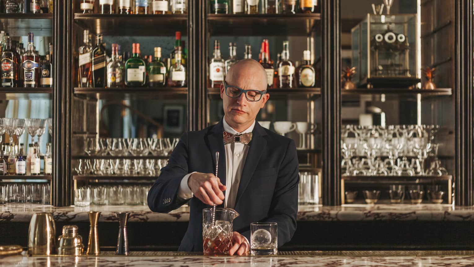 A bartender in a suit and bowtie stirring a drink with cabinets of liquor behind him.