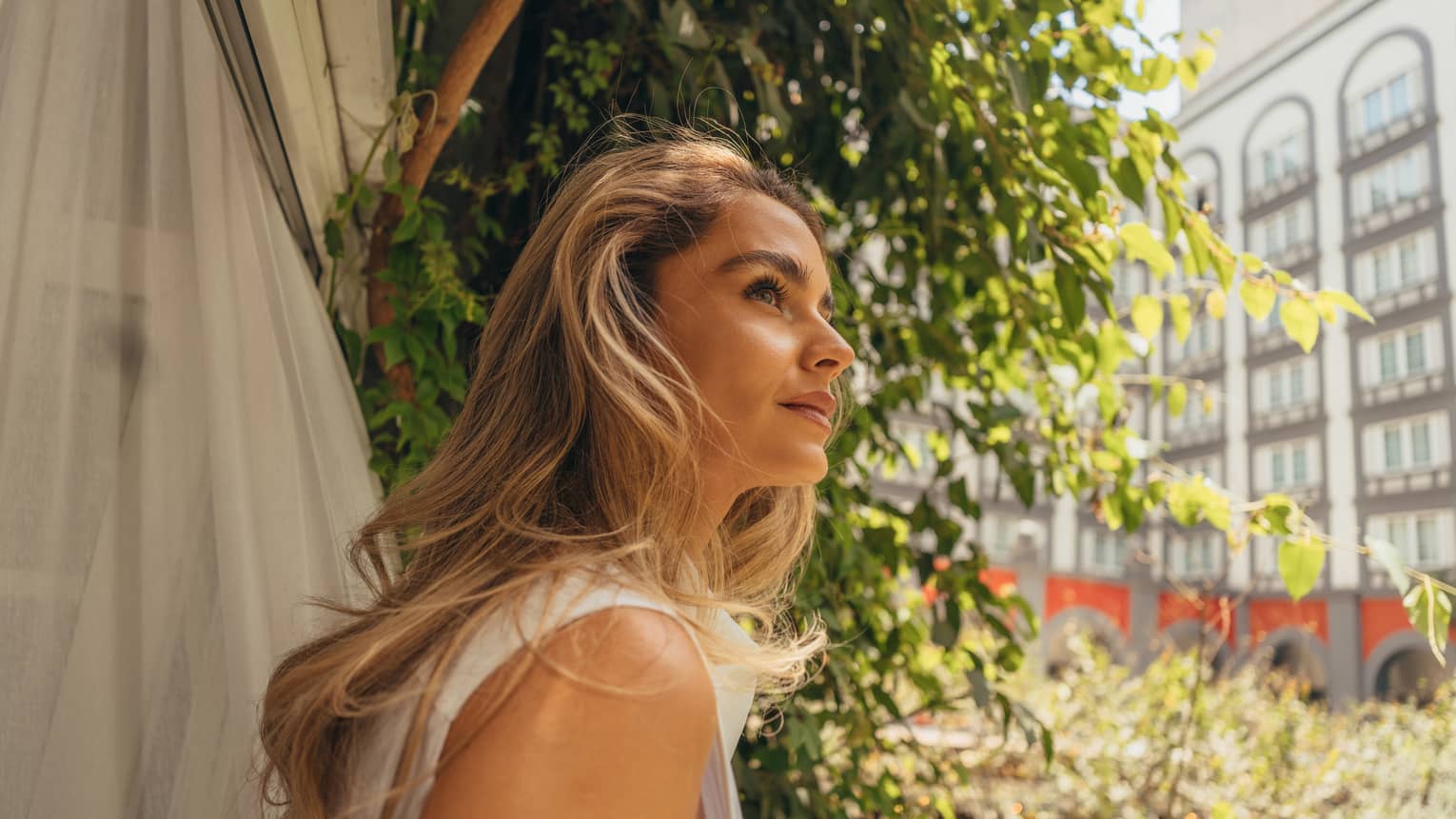 A person in an outdoor setting with foliage in the near background and the wall of a building with windows in the far background gazes off camera