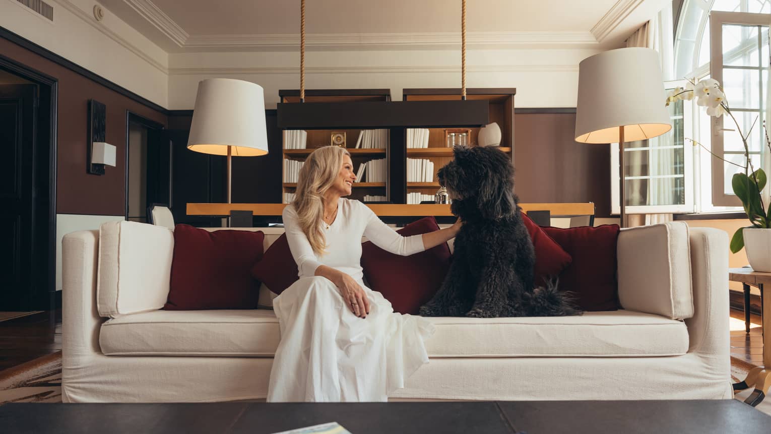 A person and a dog sit on a white couch with red pillows in a well-lit room with dark walls, a white ceiling and large windows
