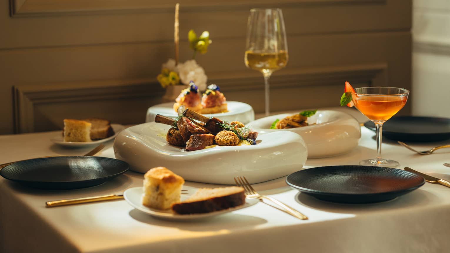 Three white serving platters with food sit in the middle of a white-cloth dining table with three black plates, a glass of white wine, a cocktail in a coupe and two small plates of bread surrounding them