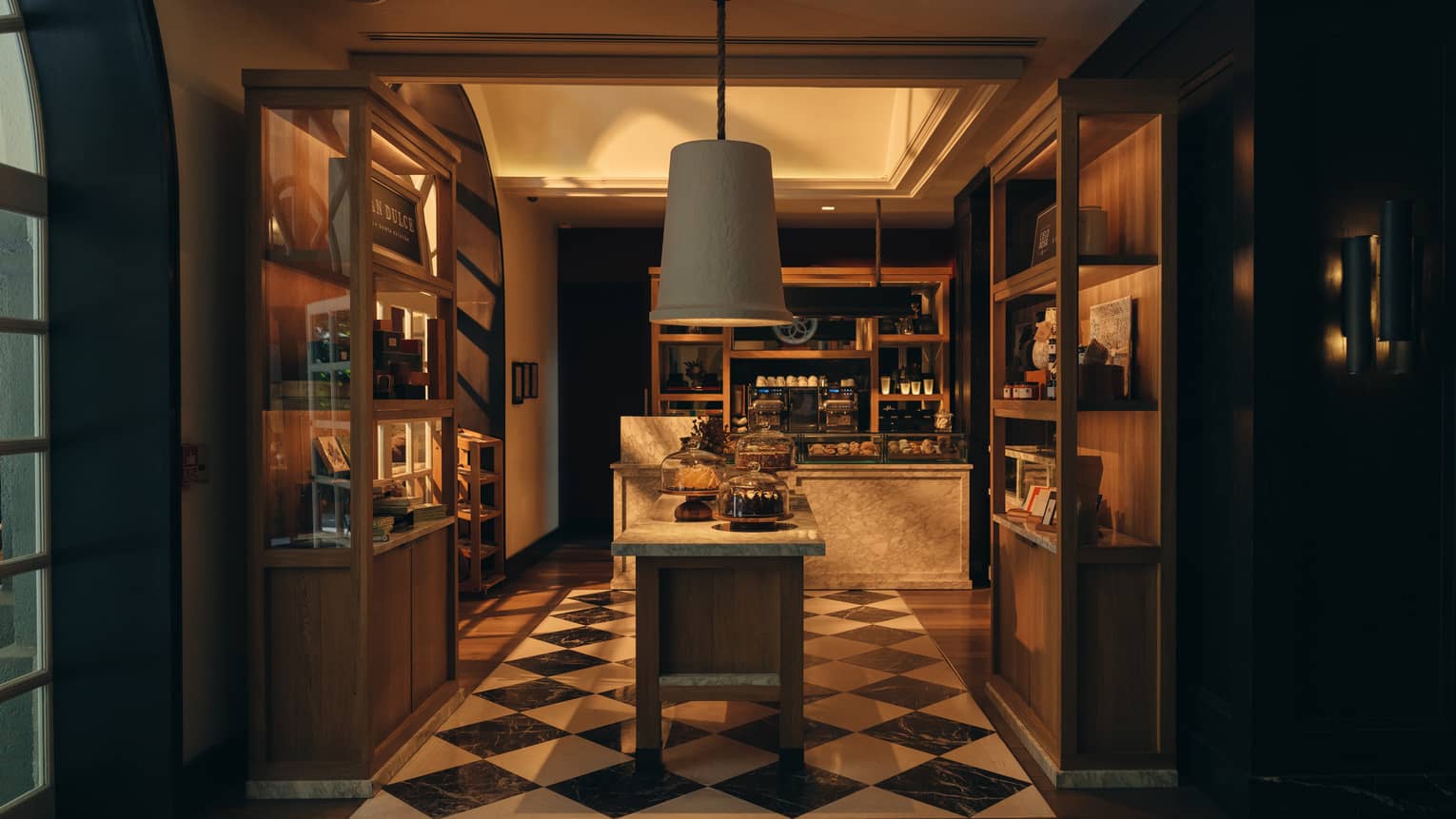Dimly lit interior of a bakery with black-and-white diamond-patterned floors, central island, wooden shelves along the side and a counter in the back