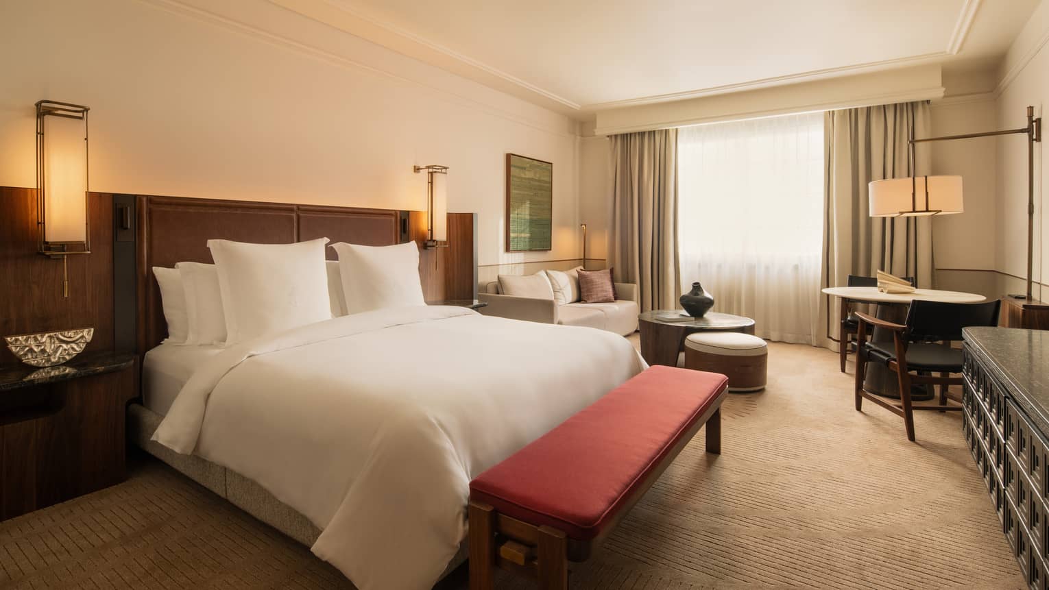 Modern hotel room featuring one king bed, red bench at the foot of the bed, lounge area with chaise and table, and window