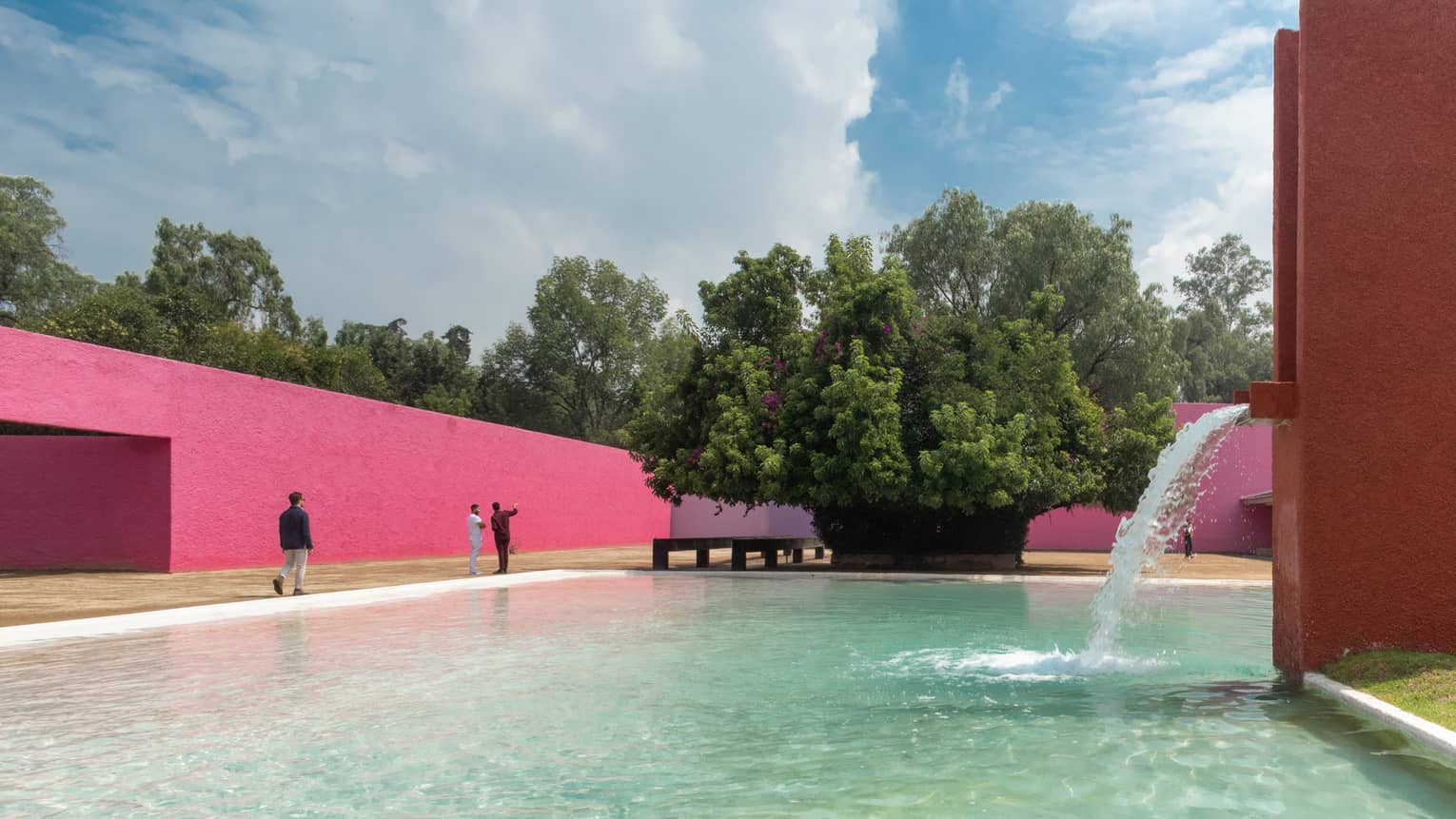 A fountain jutting out of a red oblong side wall pours water into a pool bordered by hot pink walls and a distant large tree.