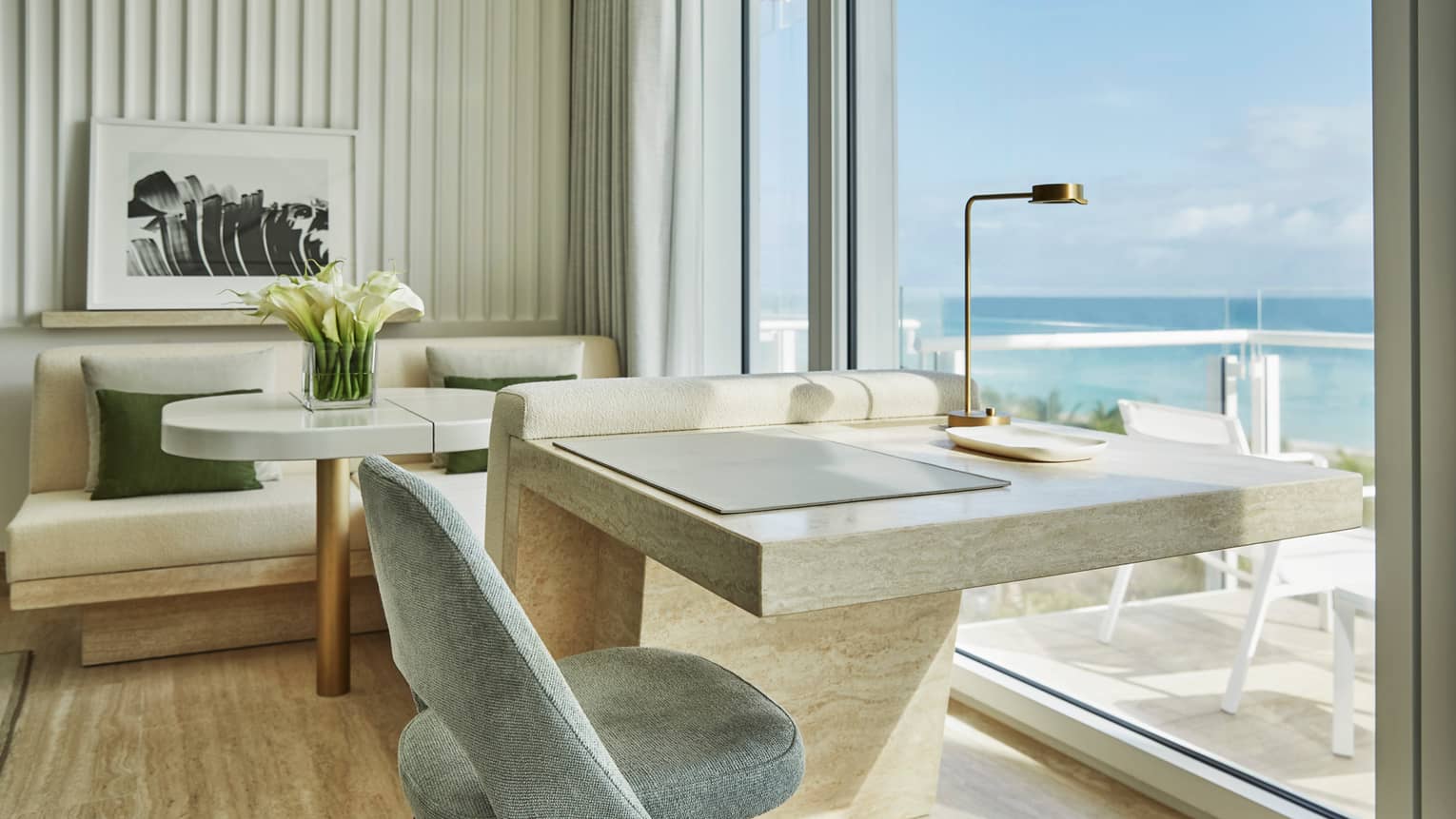 Luxury hotel room with a marble desk, a chair, a seating area with a table and flowers and floor-to-ceiling windows offering ocean views