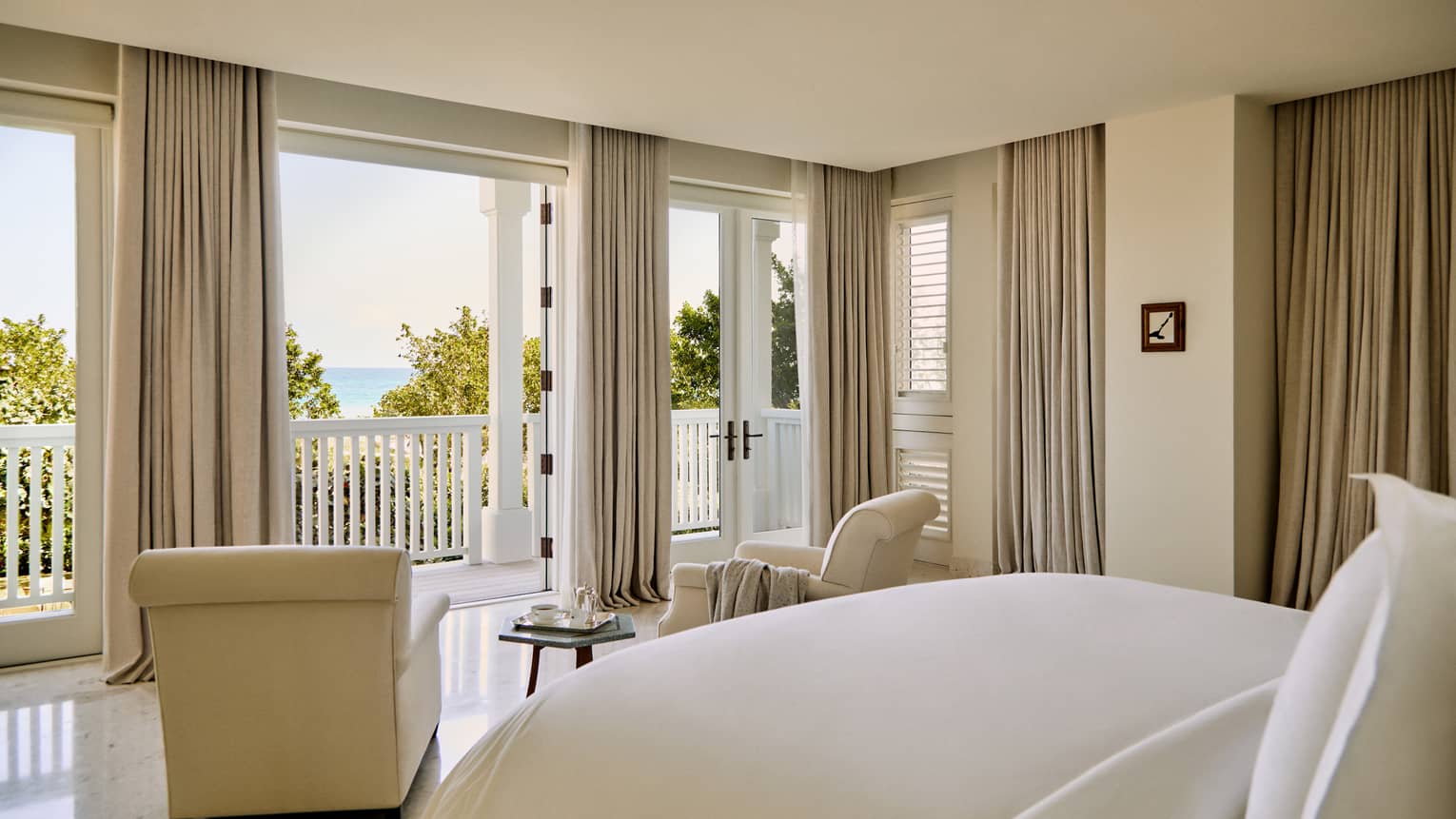Luxury villa bedroom featuring a king bed and two beige arm chairs looking out to ocean view