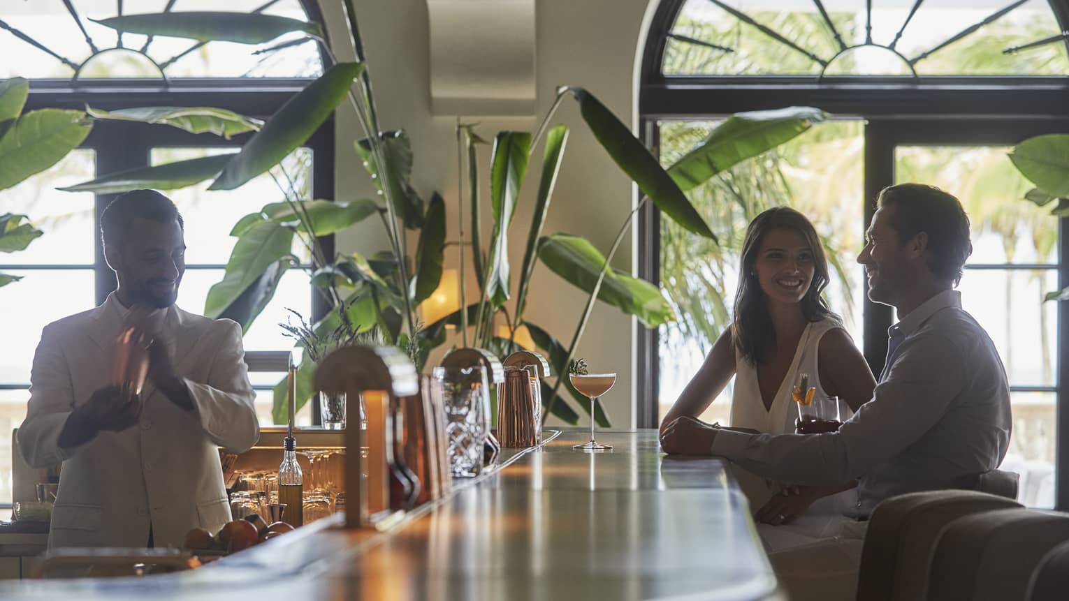 Smiling man and woman sit at bar while bartender makes their drinks 