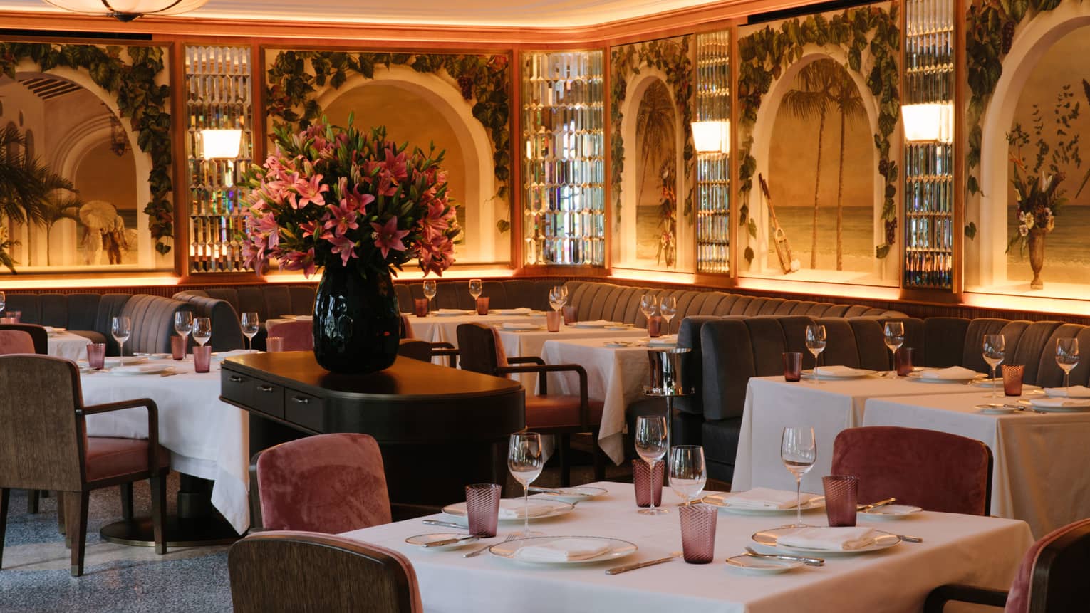 An empty dinning room of plush velvet chairs and tropical inspired murals. A large bouquet of pink hibiscus flowers sits in the middle