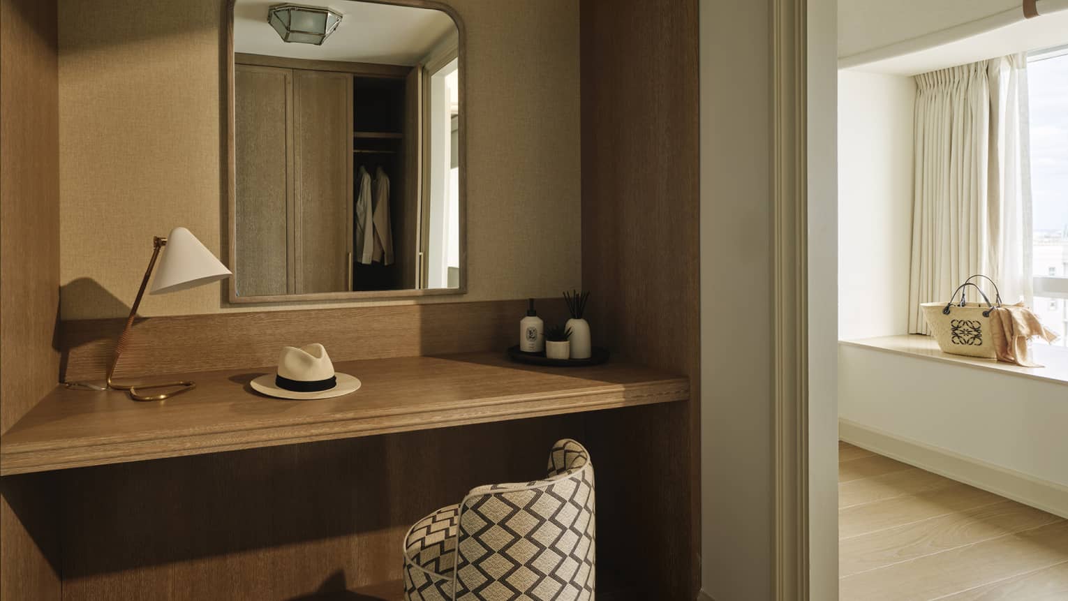 Hotel suite bedroom with a built-in wooden vanity area, featuring a mirror, lamp and upholstered stool