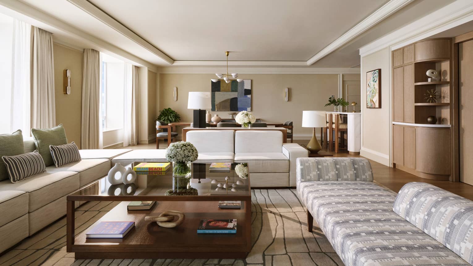 A hotel suite's living room with two off-white sofas and a patterned chaise surrounding a large square wooden coffee table